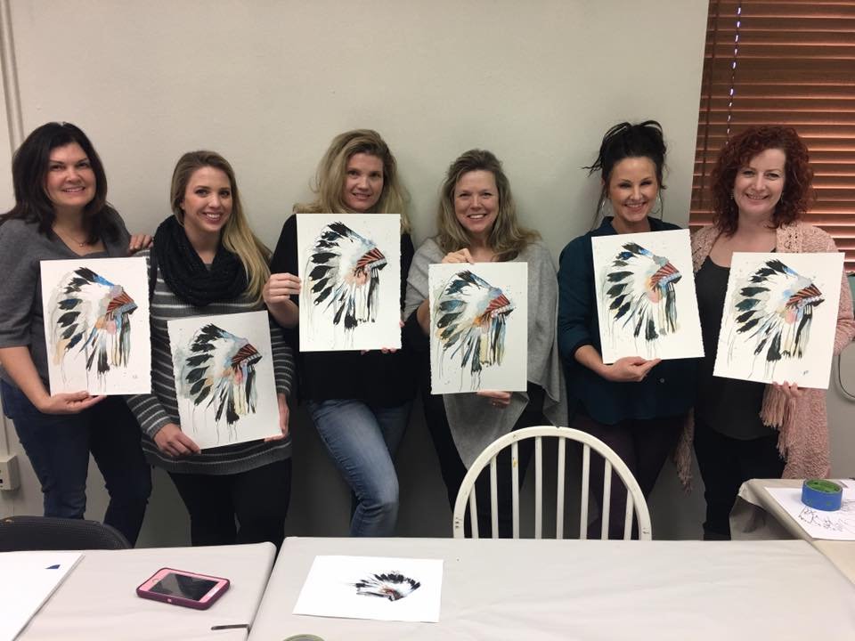 Six women standing inside holding paintings of a Native American headdress, smiling at the camera.