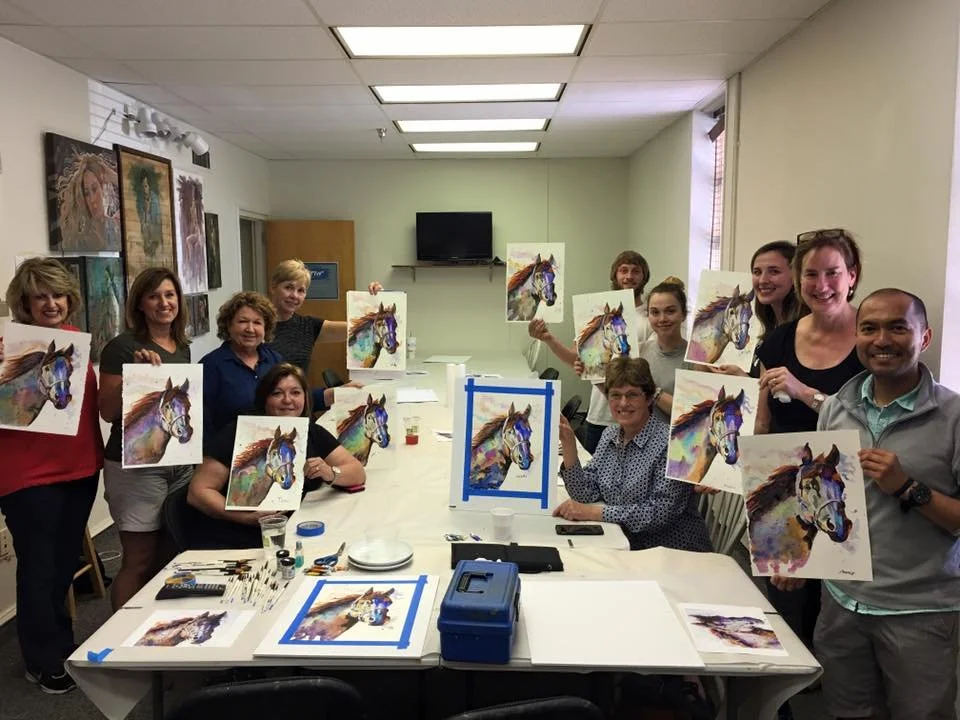 A group of people in a room holding up watercolor paintings of a colorful horse. They are gathered around a table with art supplies, and everyone is smiling.