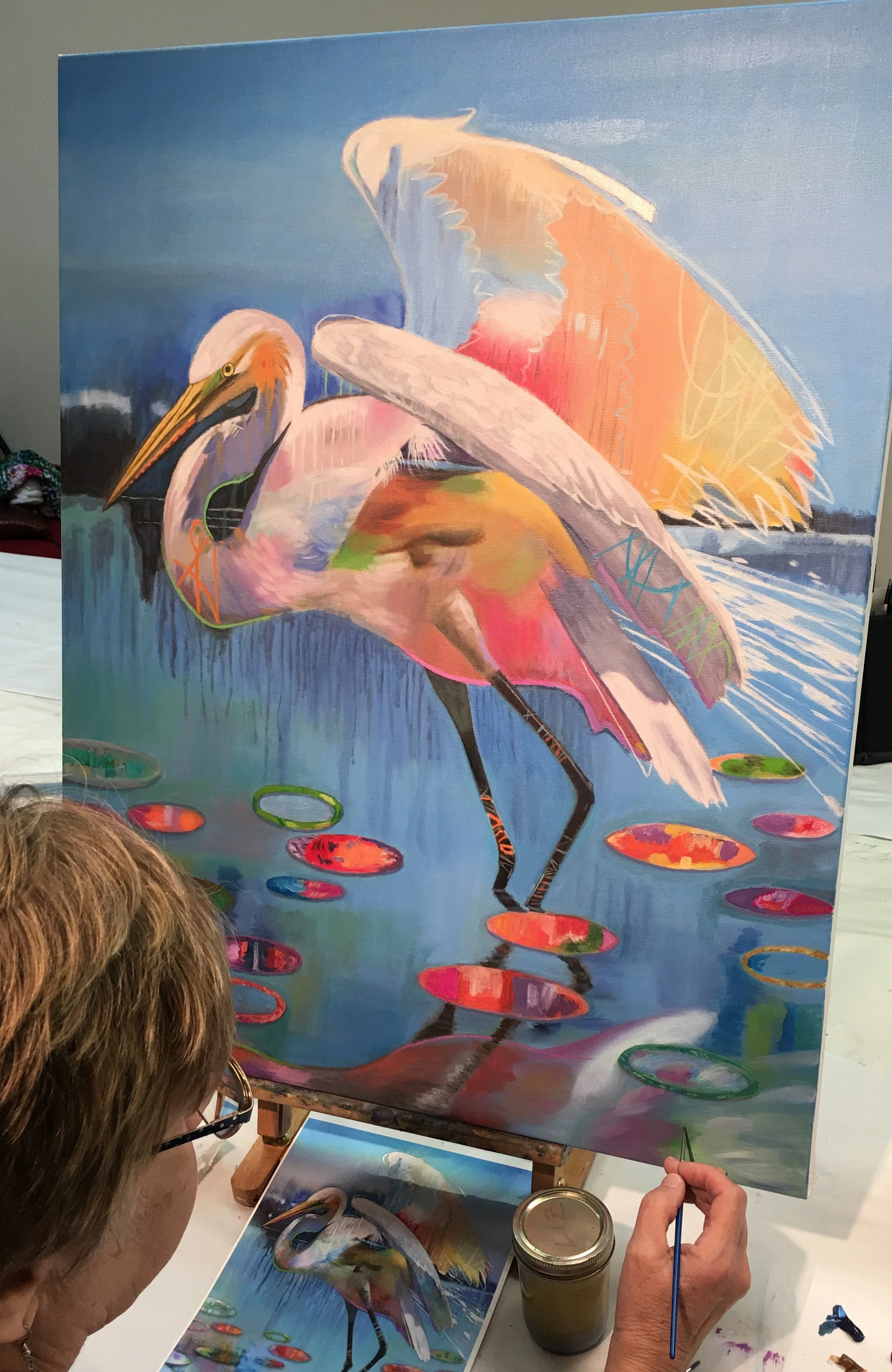 An artist painting a colorful, abstract depiction of an egret standing in water with lily pads, with a reference photo on the table.