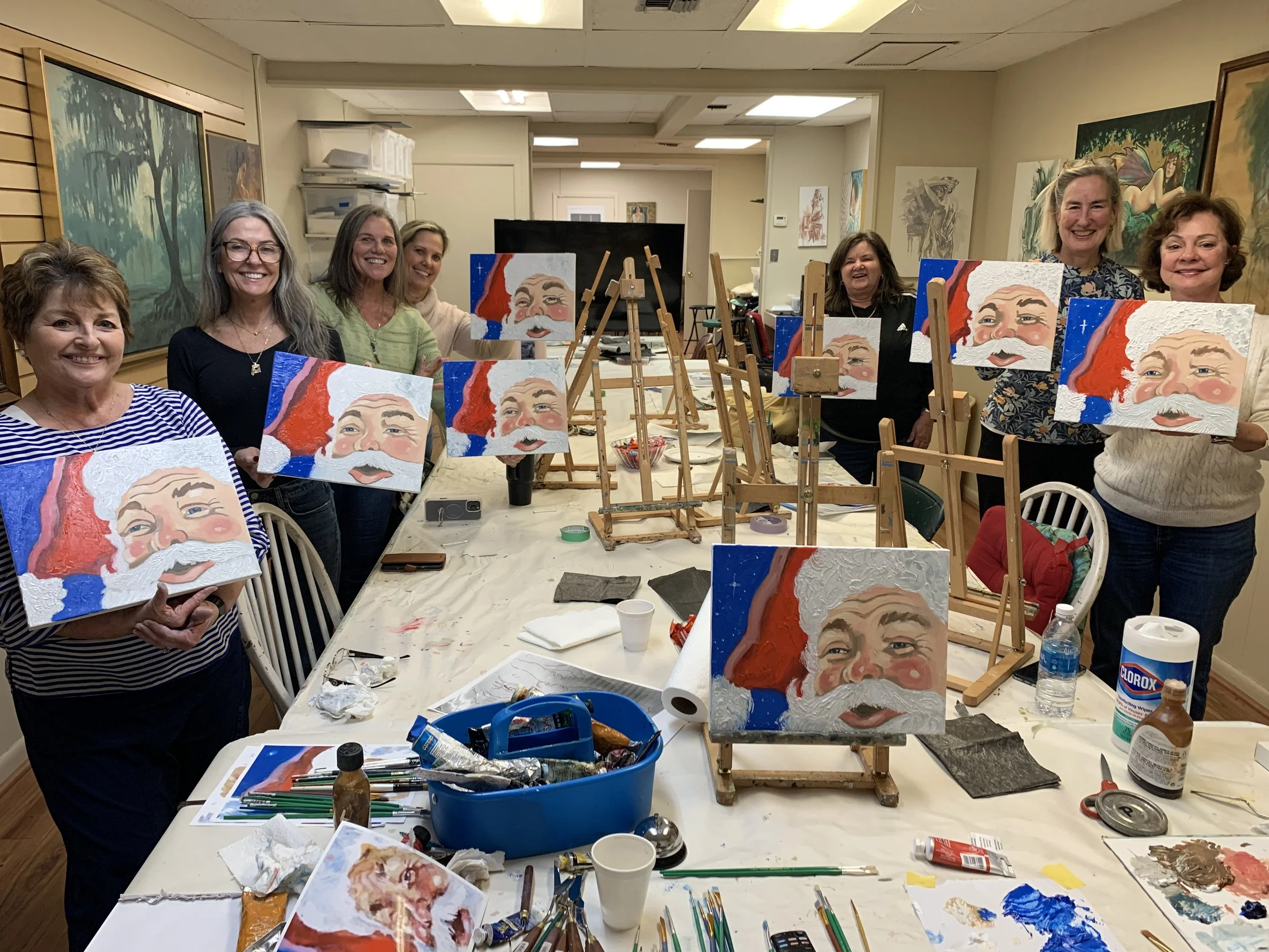 Group of women at a painting class holding up completed Santa Claus portraits, with easels and painting supplies on the table.