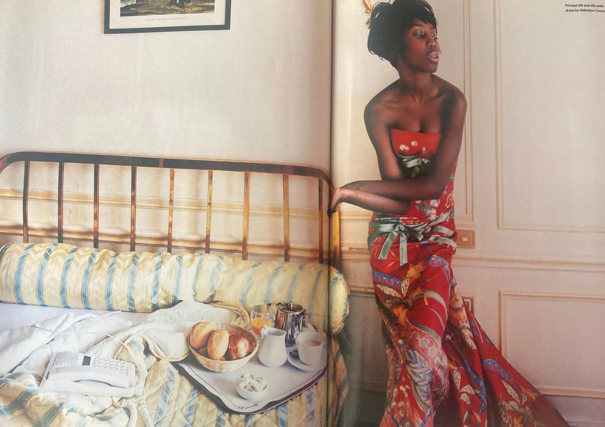 Double page fashion editorial from ELLE UK December 2003 showing a black woman with short hair wearing a red printed floor length dress on the right and a bed with various breakfast items on the left.