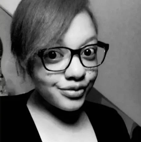 Image shows a black and white photo of a black woman with a side fringe wearing thick black framed glasses and a black t-shirt