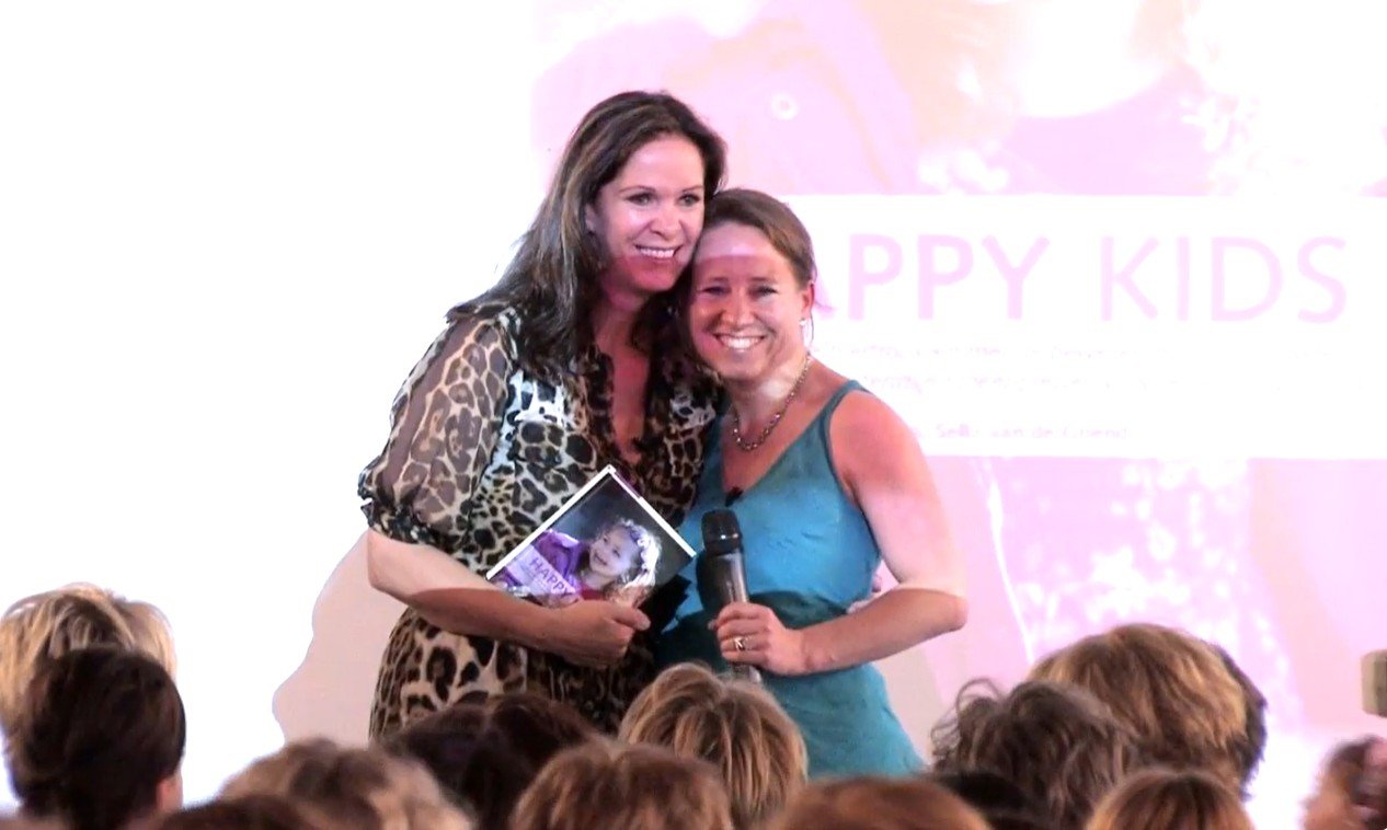 Twee vrouwen op een podium omhelzen elkaar, één houdt een foto vast en de ander houdt een microfoon. Op de achtergrond is een banner met de tekst 'HAPPY KIDS' zichtbaar. Er is een publiek voor het podium.