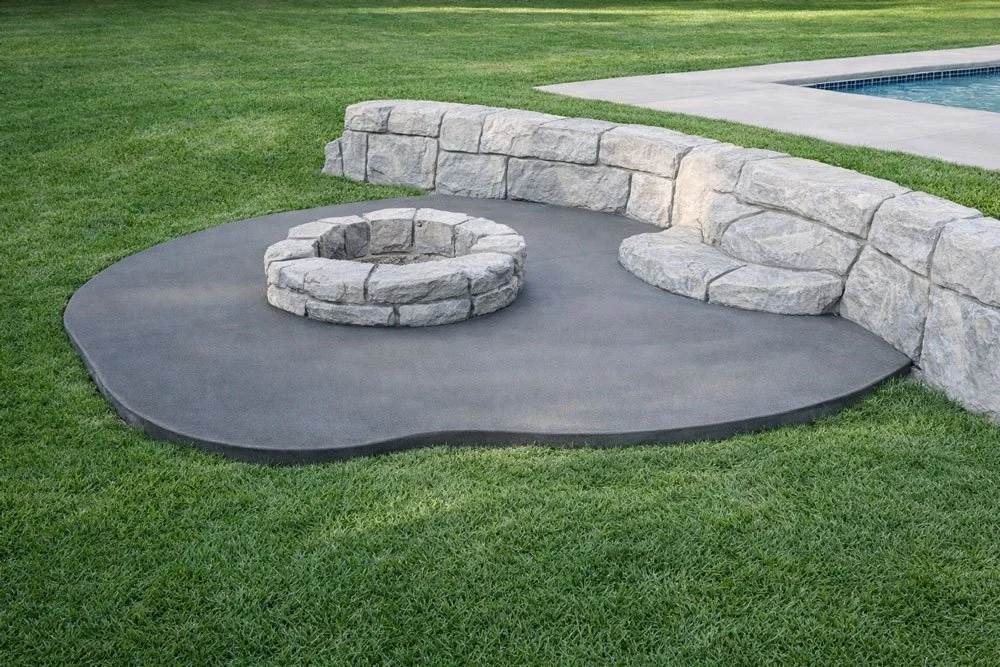 A backyard with a stone fire pit, surrounded by a curved stone wall, on a concrete patio area next to a grassy yard and a swimming pool.