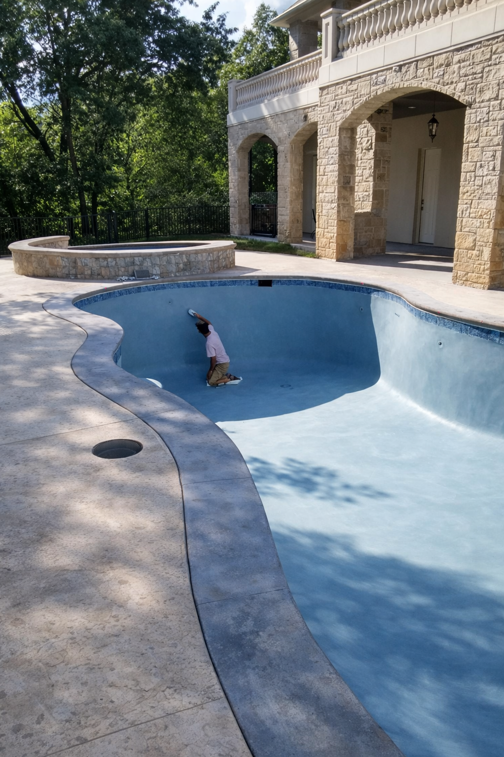 Man in a pink shirt and tan shorts cleaning an empty swimming pool with a brush, with a house and trees in the background.