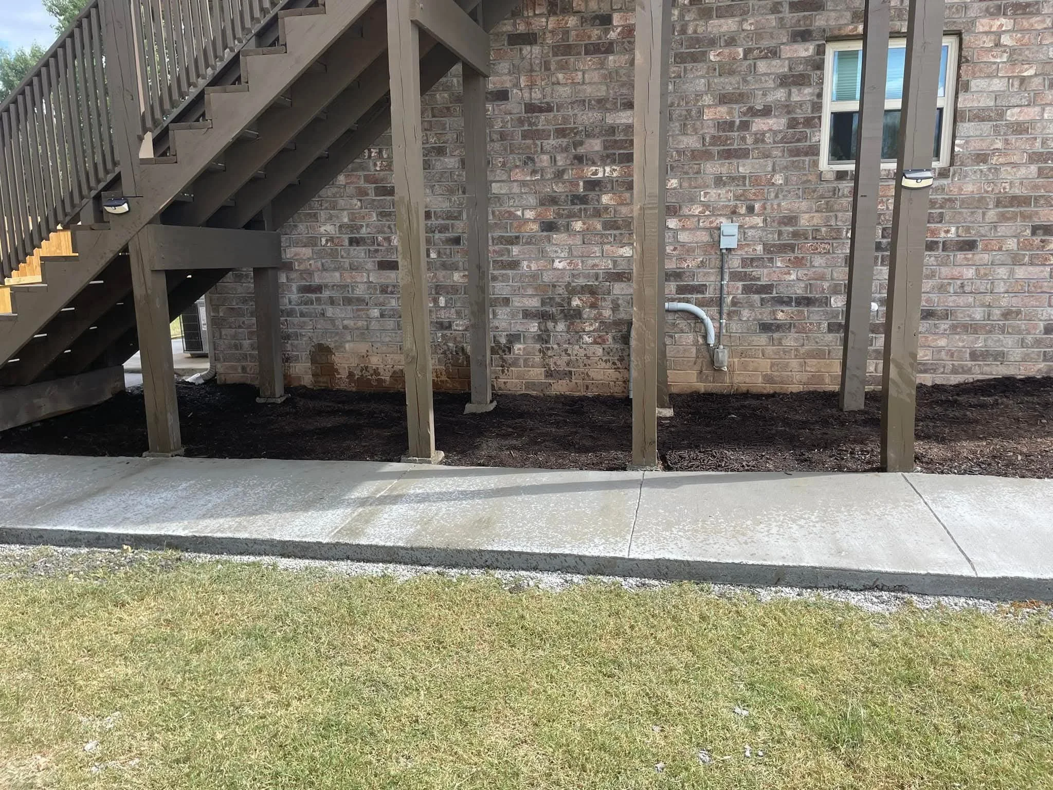 Backyard with brick house wall, wooden stairs leading up, green grass, concrete patio, and electrical wiring on the wall.
