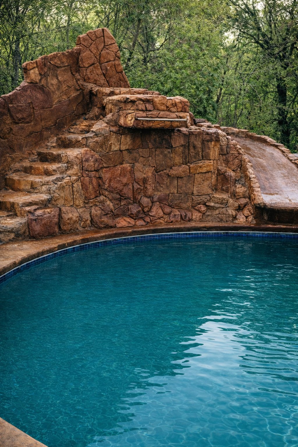 A swimming pool with a rock waterfall feature and steps surrounded by greenery.