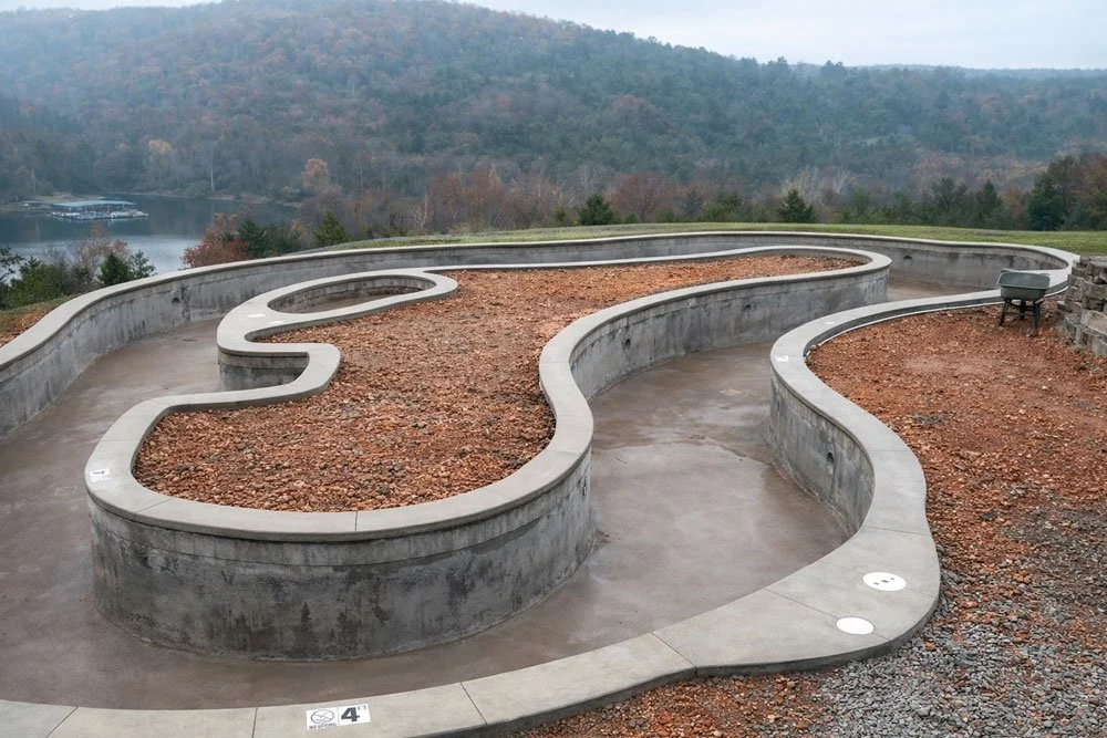A winding concrete walkway on a hilltop with a view of trees, a lake, and mountains in the background. The pathway has a curvy design with some sections filled with reddish-brown gravel.