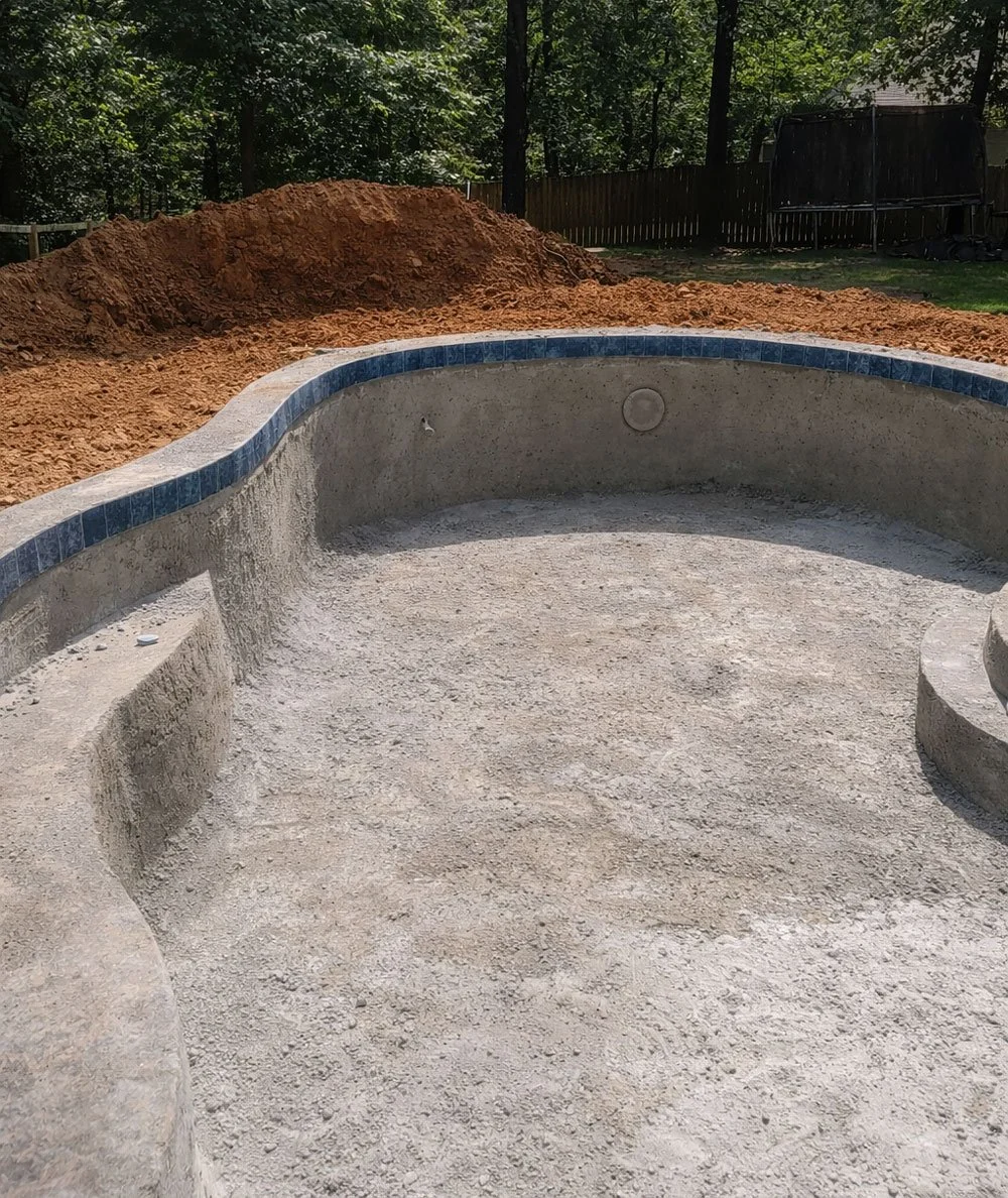 Empty in-ground swimming pool under construction with a dirt mound behind it, surrounded by a backyard with a wooden fence and trees.