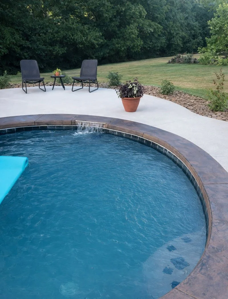 A backyard scene with a swimming pool in the foreground, surrounded by a concrete patio. There are two black chairs with a small table between them on the patio, with a decorative item on the table. A large potted plant is nearby. The background features a grassy lawn bordered by trees and bushes.