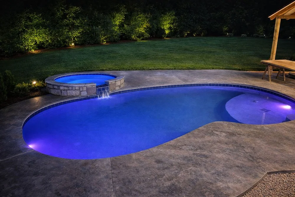Nighttime view of a backyard with a lit swimming pool and spa, surrounded by a stone patio, a grassy lawn, and trees in the background.