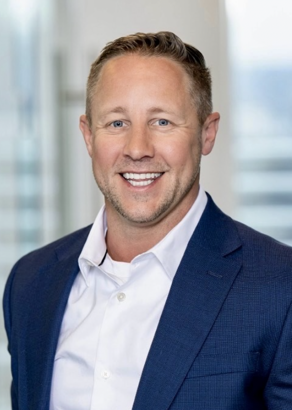 Headshot of a smiling man in a white shirt and dark blazer, in an office setting with bright windows in the background.