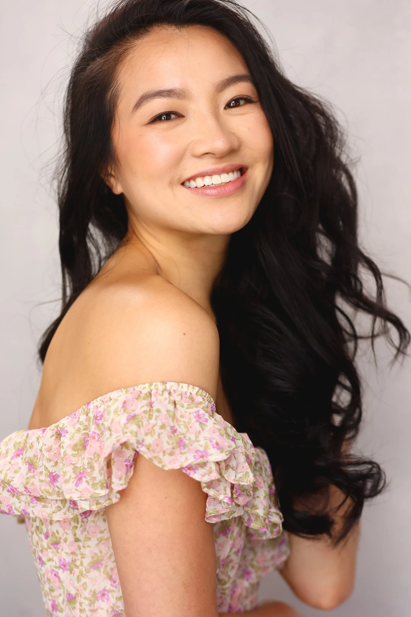 A young woman with long, curly black hair smiling and looking at the camera. She is wearing a floral off-the-shoulder top and standing against a plain light background.