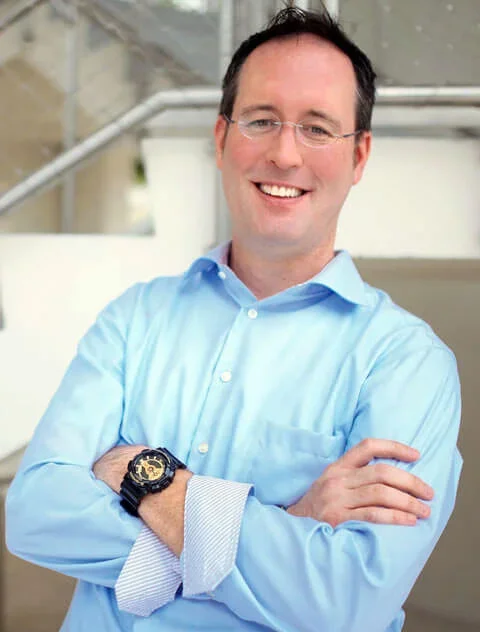 Smiling man wearing glasses and a light blue dress shirt, crossing his arms, standing outdoors with a modern building background.