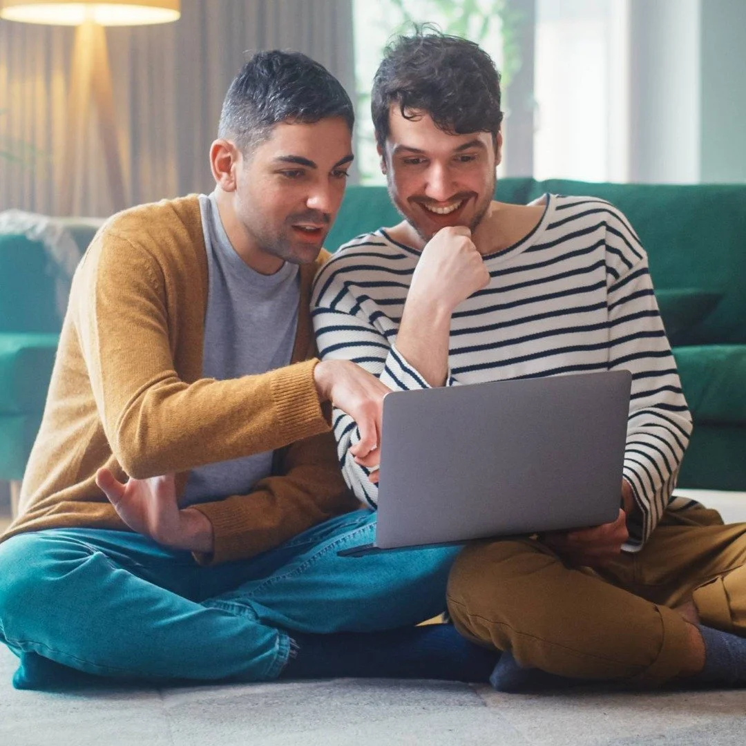 Two young people sitting comfortably on the carpet, looking at a laptop together and smiling while online with their therapist.