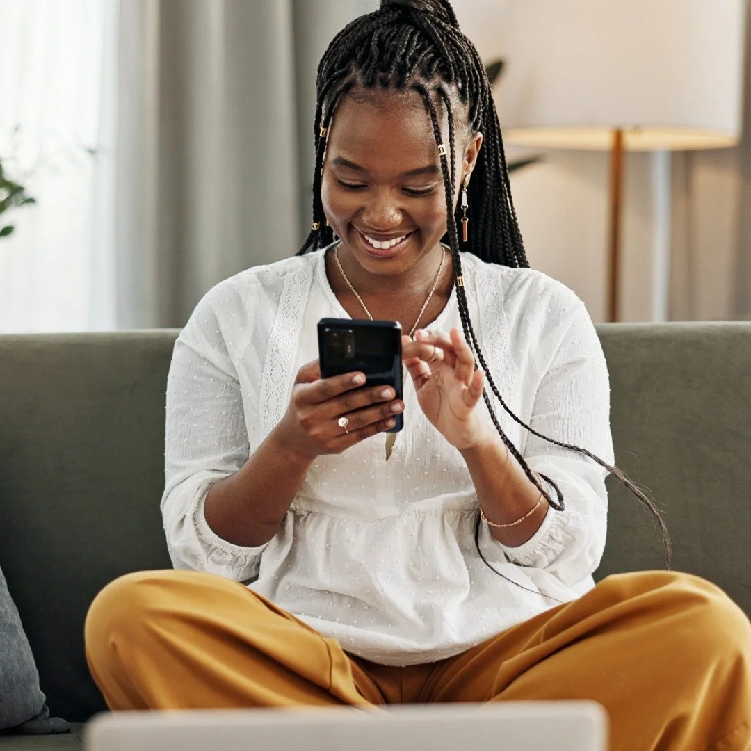 A smiling woman with braided hair, wearing a white blouse and mustard-colored pants, sitting on a couch and looking at her phone.