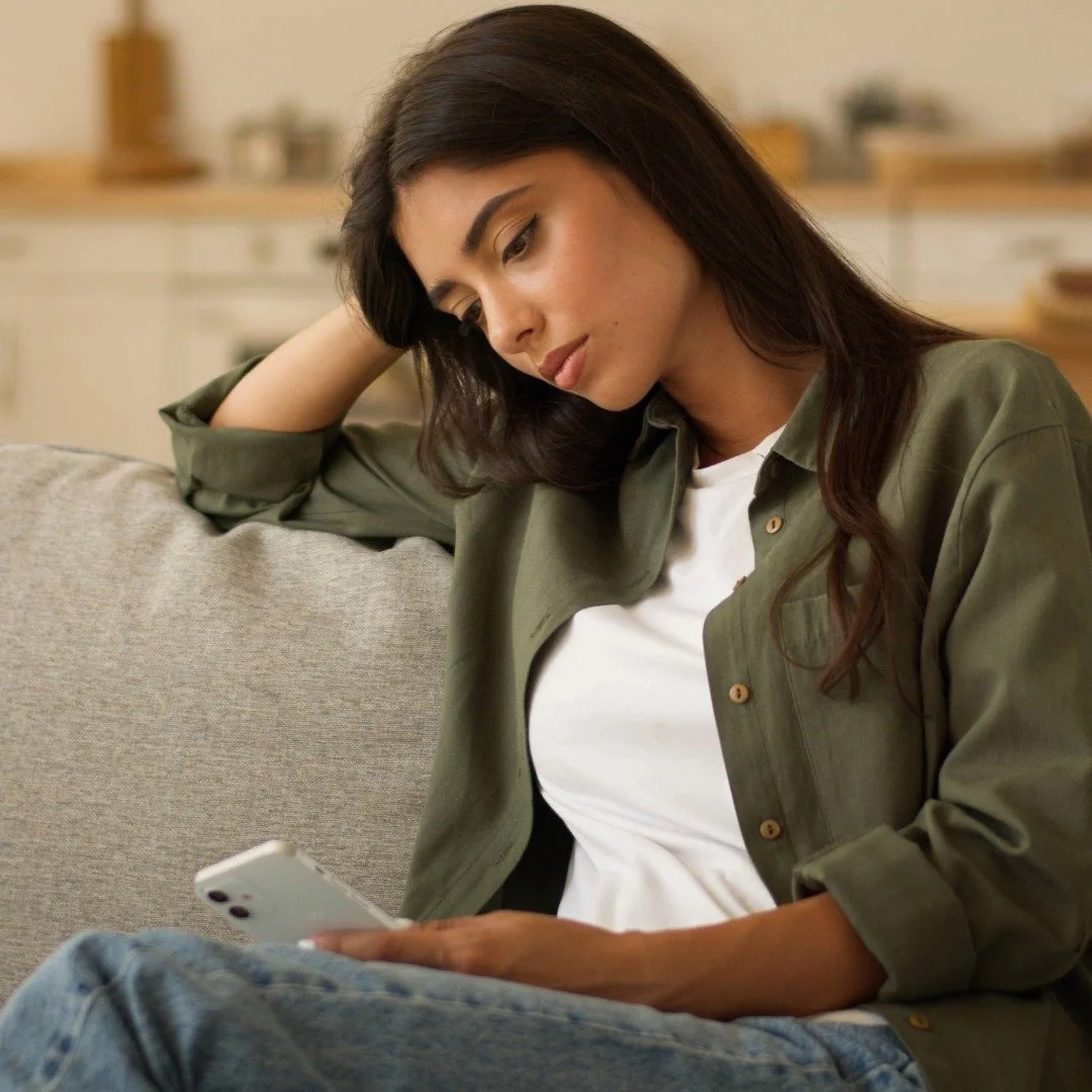 A young woman sitting on a couch, looking at her phone with a thoughtful expression, while talking with her therapist.