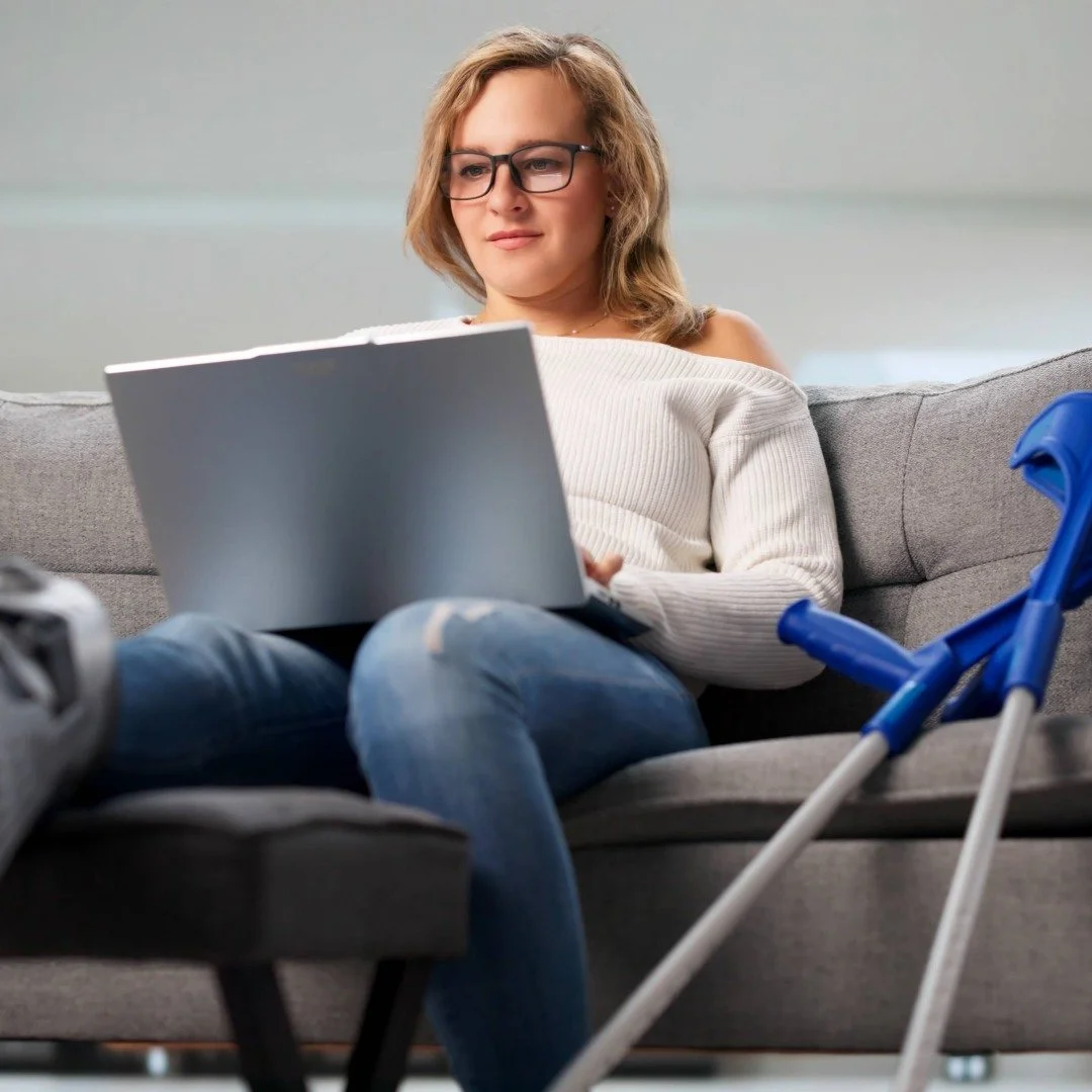A young woman sitting on a couch, with a laptop and crutches leaning against the couch beside her.