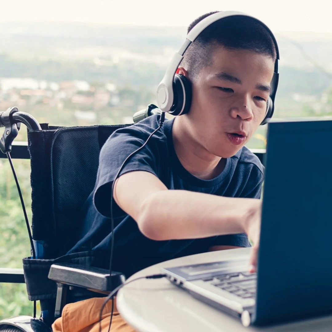 A young person with a headset using a laptop for their virtual therapy session, sitting in a wheelchair with a view of a city landscape in the background.