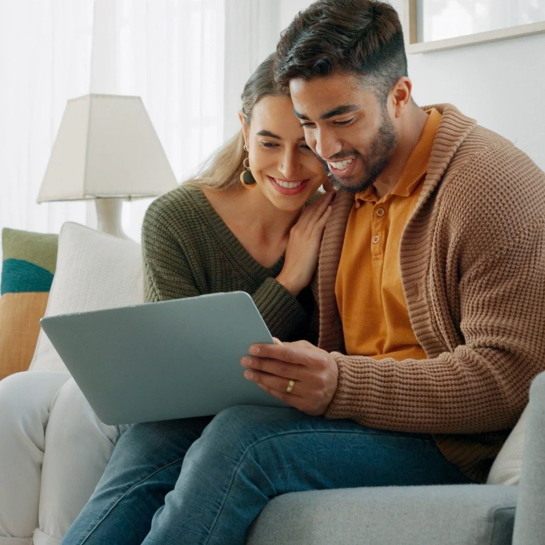A happy couple sitting on a sofa, looking at a laptop for their virtual therapy session.