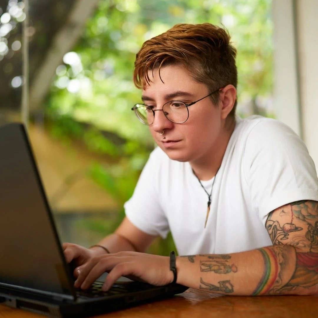 Young person with short brown hair, glasses, and tattoos on their arms working on a laptop at a wooden table outdoors with green trees in the background.