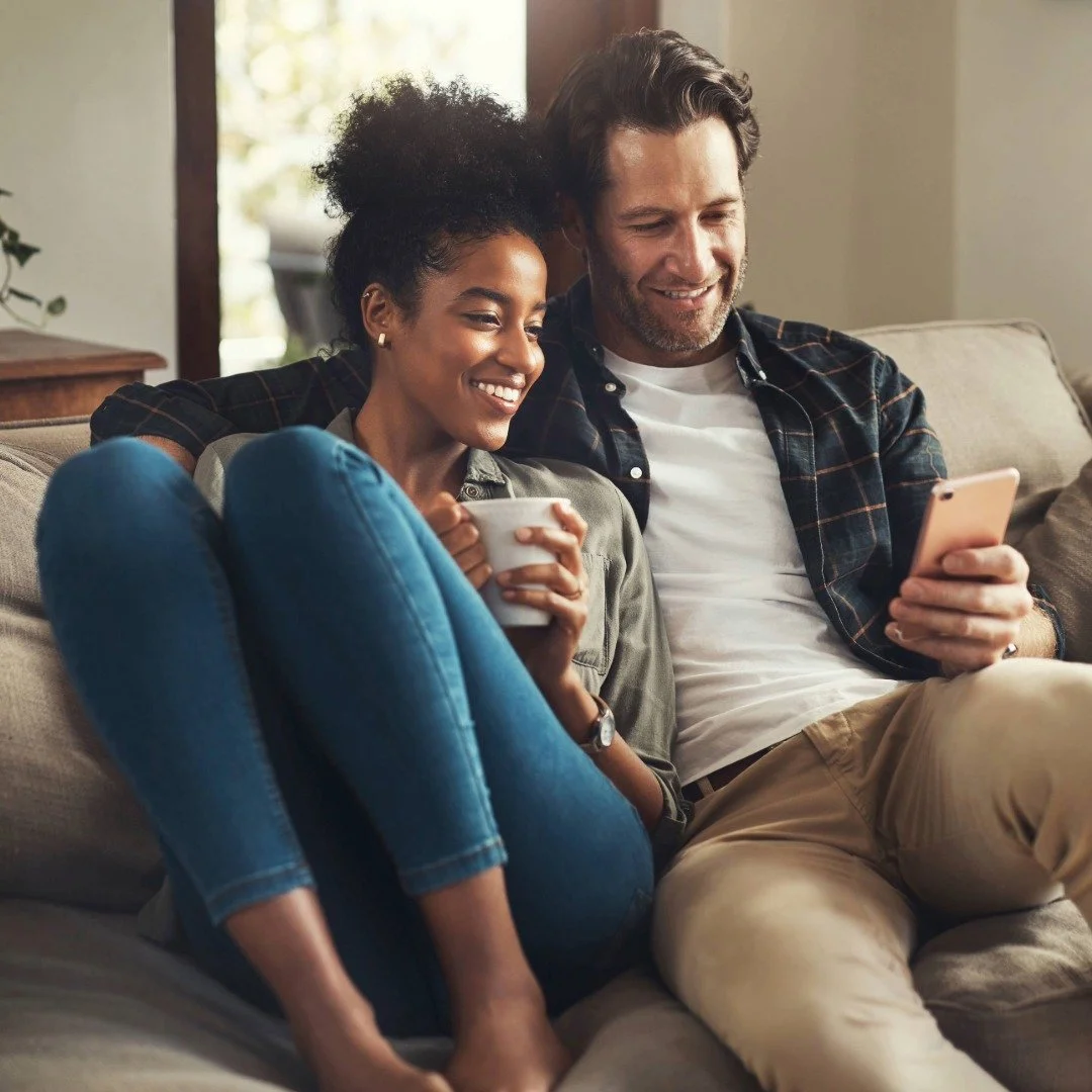 A couple snuggled on a couch, smiling and looking at a smartphone together, enagaged in a phone therapy session.