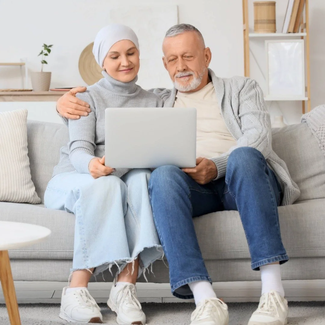 A couple sitting on a sofa at home, looking at a laptop together while having a virtual meeting with their therapist.