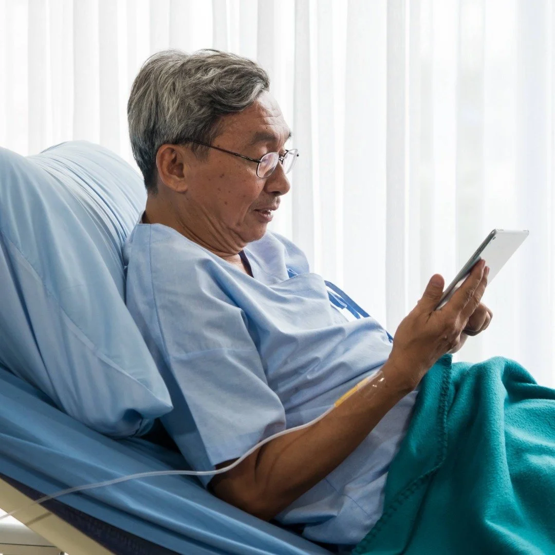 An elderly man in a hospital bed wearing glasses, looking at a tablet while receiving online care from a therapist.