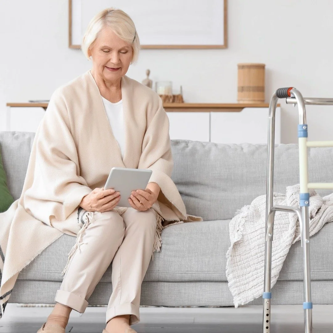 An elderly person sitting on a light gray sofa, talking to a therapist on her tablet, with a walker beside her.