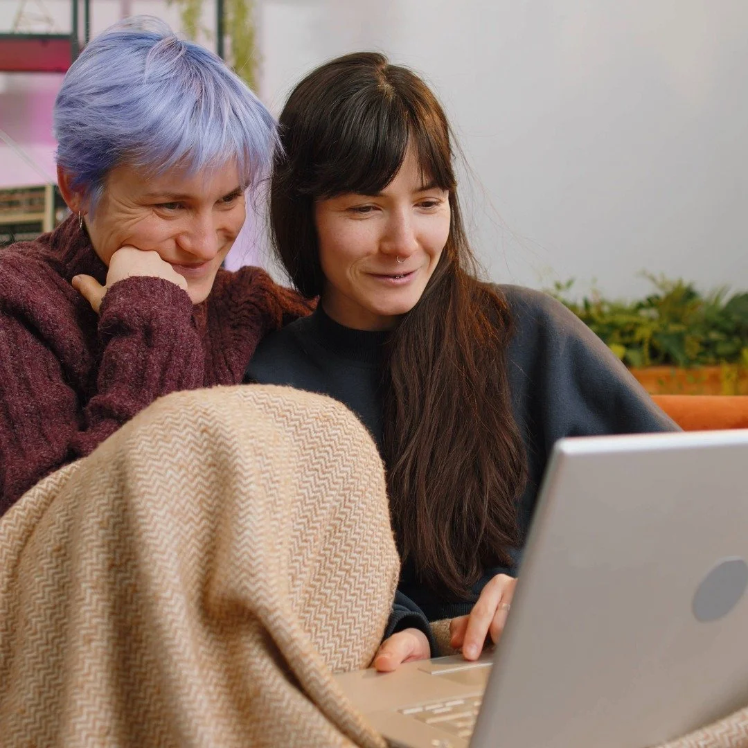 Two people sitting closely together looking at a laptop screen, smiling, in a cozy indoor setting while on a virtual therapy session.