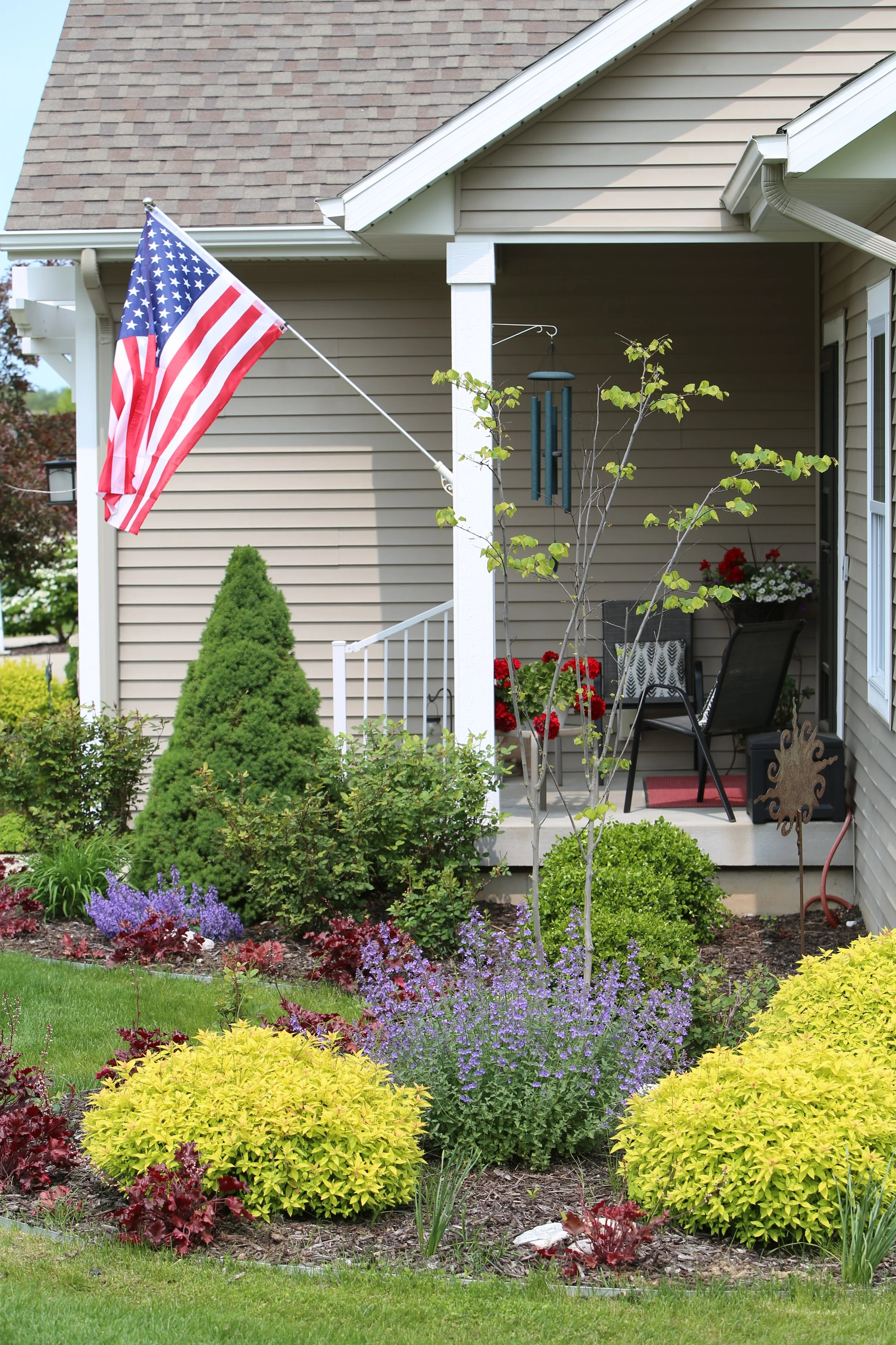 A front porch decorated with potted red flowers, a wind chime, and outdoor furniture, with a garden of colorful bushes and a small tree in front and an American flag mounted on the house.