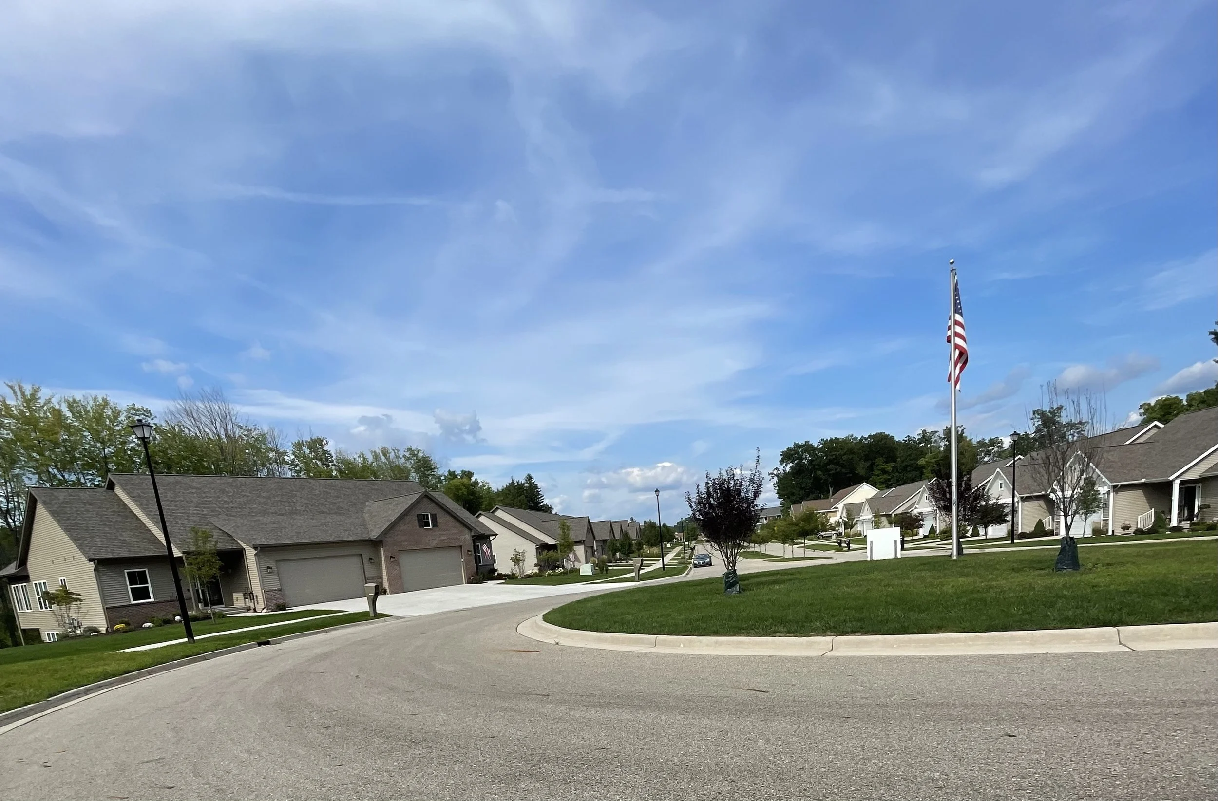 Residential neighborhood with single-family homes, a flagpole with an American flag, a clear blue sky with some clouds, and a curved street with green grass and trees.