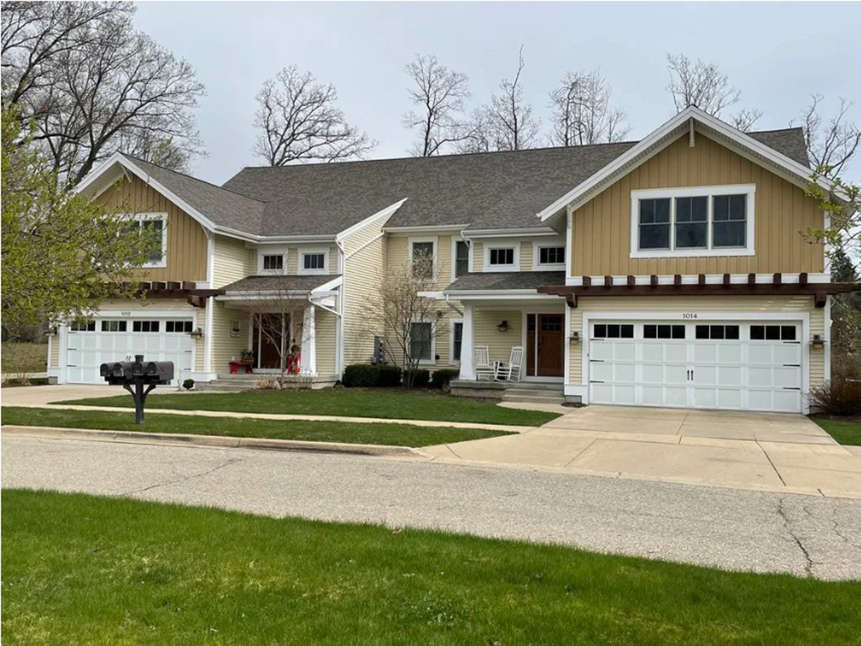 A two-story house with a yellow and beige exterior, white garage doors, a small front porch with white chairs, and a black mailbox in front.