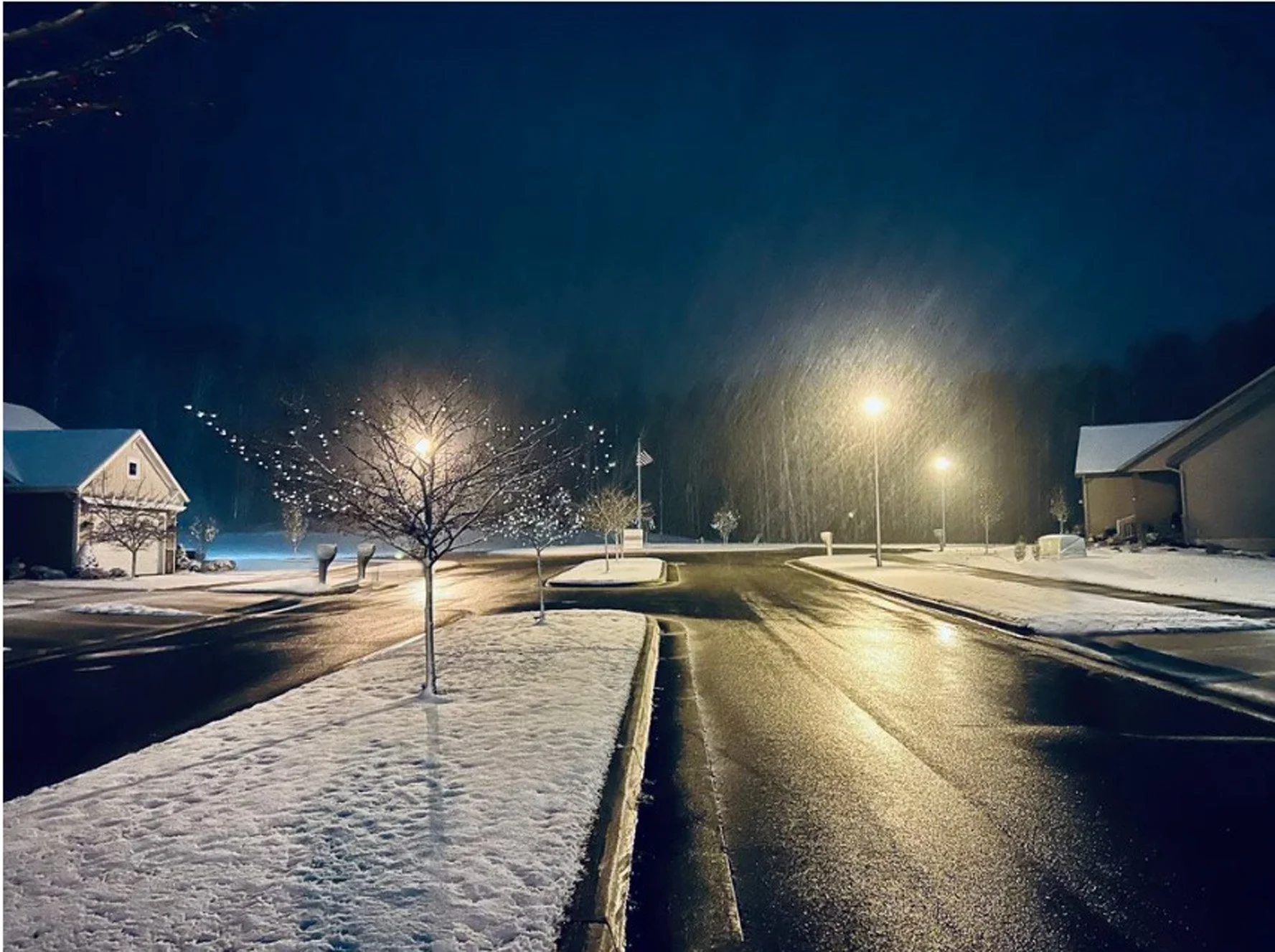 A snowy neighborhood street at night with wet asphalt reflecting streetlights, snow on the lawns and rooftops, and a few small trees lining the sidewalk.