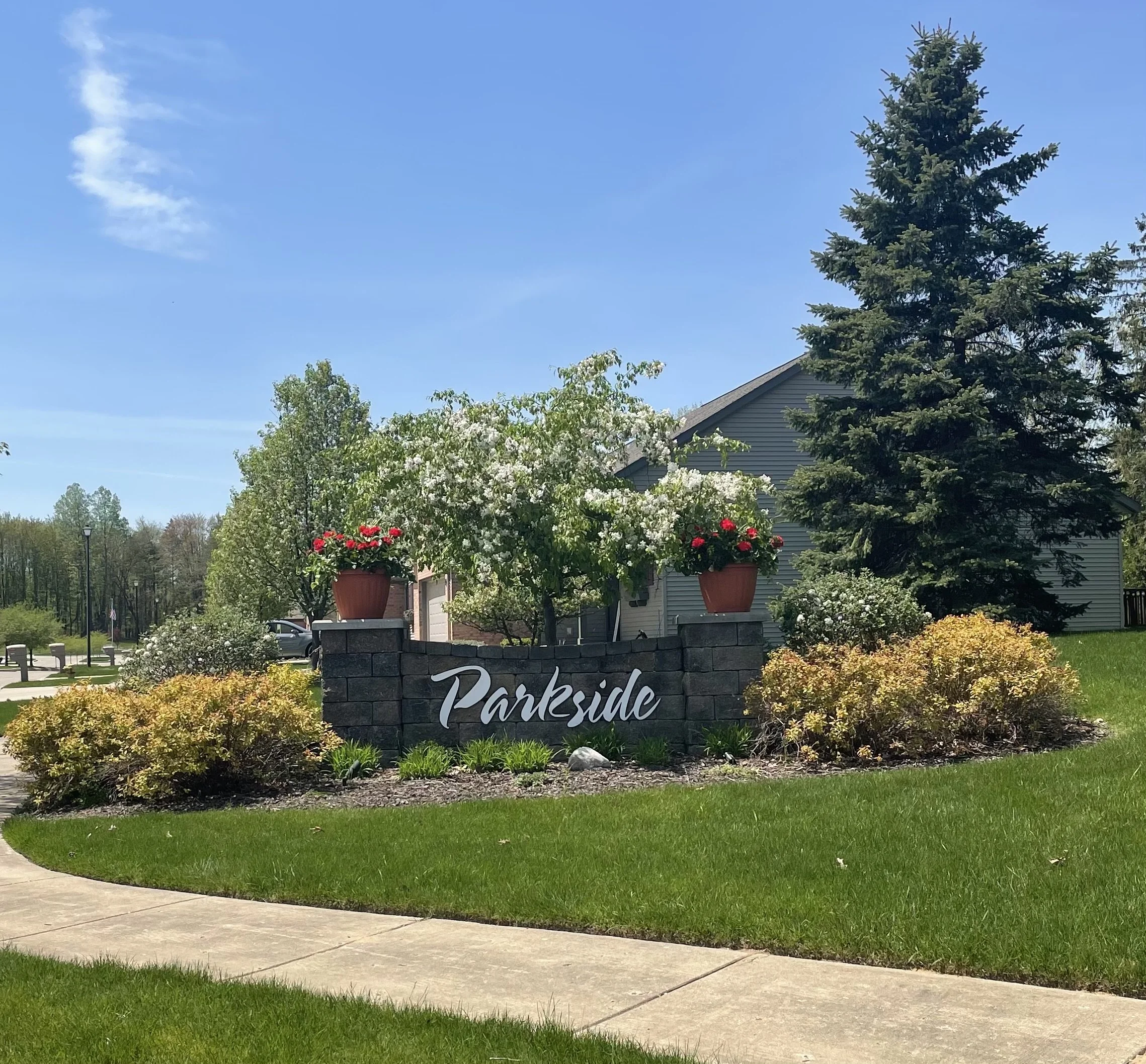 Entrance sign reading 'Parkside' with decorative plants and large flower pots, surrounded by a well-manicured lawn and trees in a suburban neighborhood