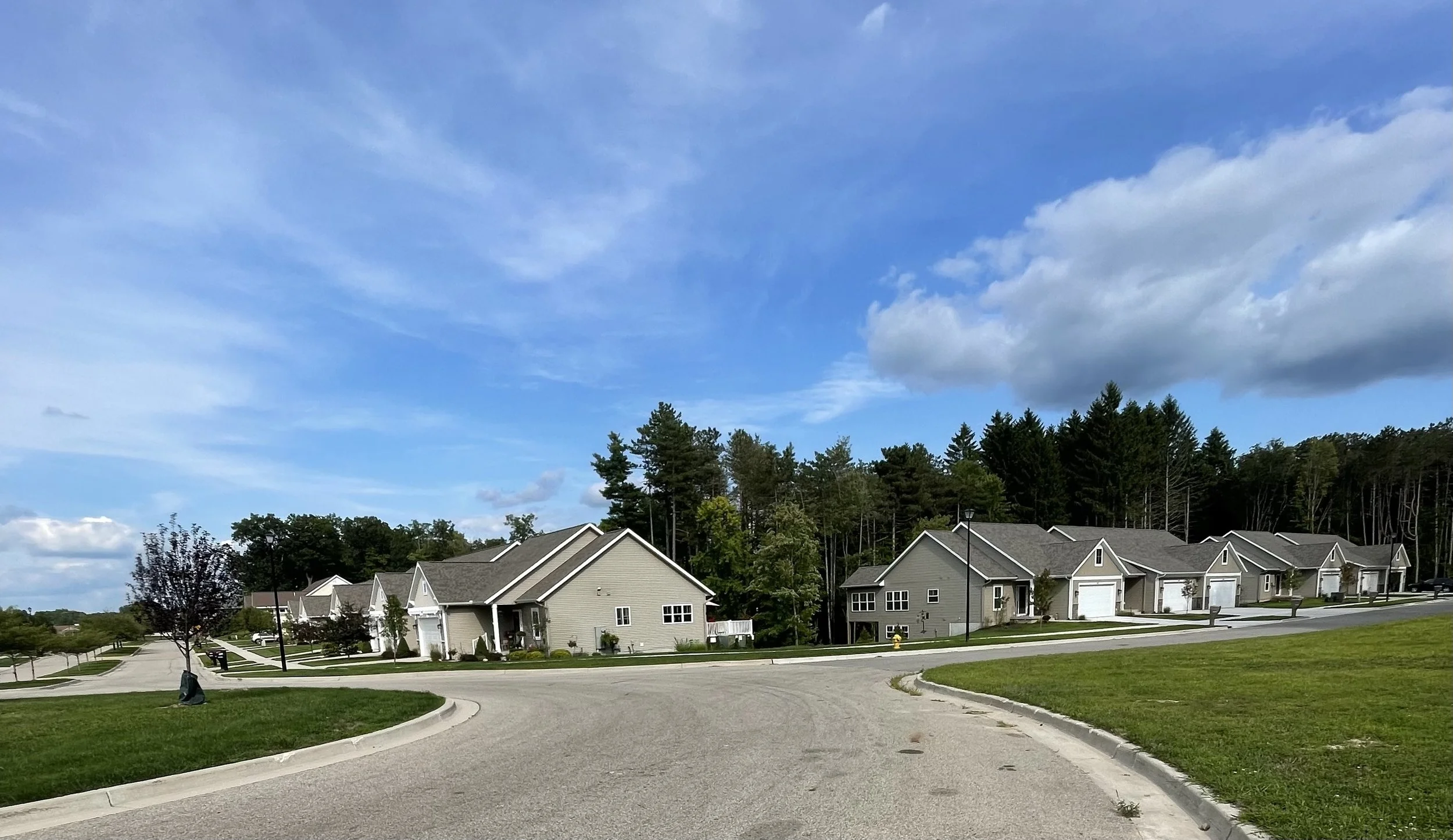 A suburban neighborhood with several modern single-family houses, well-maintained lawns, and a driveway in a curved street. Trees and a blue sky with some clouds are in the background.