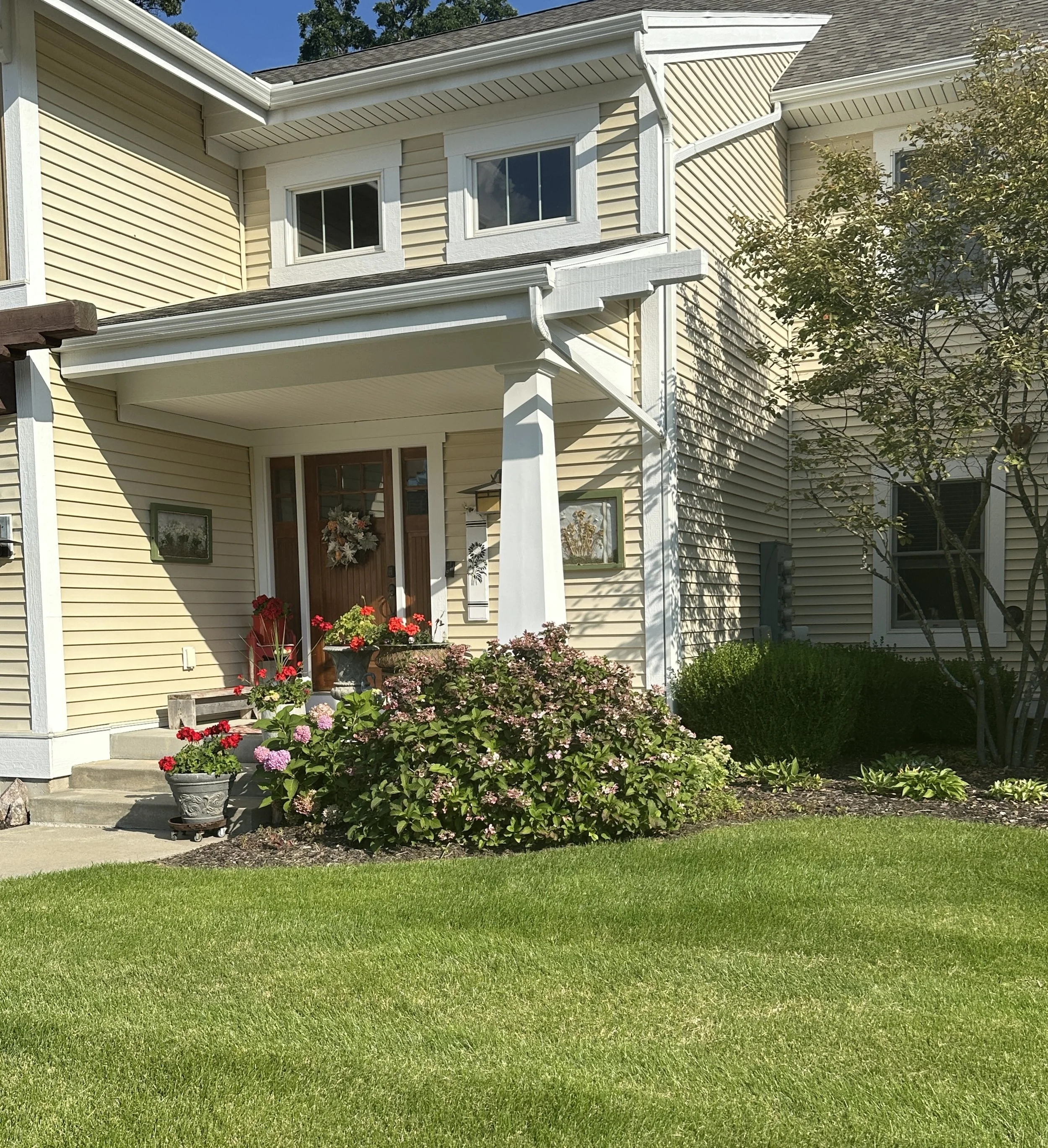 Front yard of a beige house with white trim, decorated with flowers and shrubs, green lawn in foreground, and a tree on the right side.