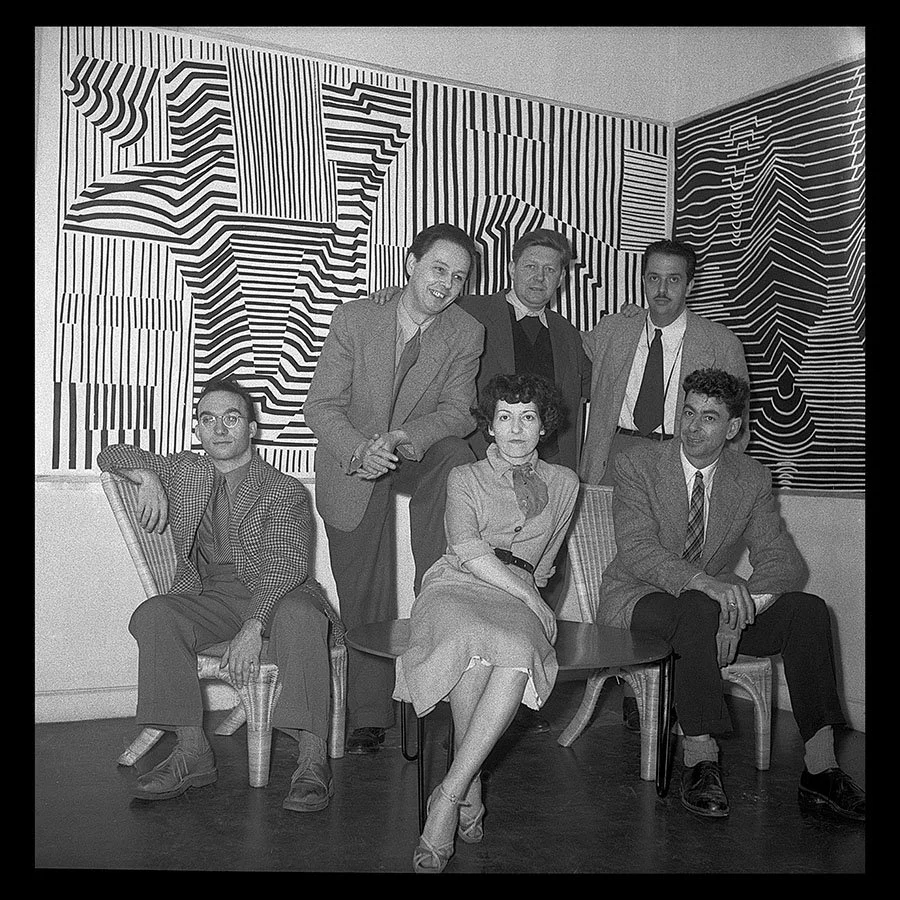 Black and white photo of six people, five men and one woman, posing in front of an abstract, geometric patterned wall. Four people are seated on chairs, and two are standing behind them. Denise René Galerie.
