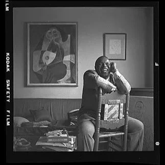 A man sits on a chair in a living room, smiling with one hand resting on his knee and the other on the back of the chair. The room has framed artwork on the wall, including a large painting of a woman with glasses and a smaller picture. There are boo