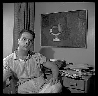 A man with a mustache sitting in a room with a desk, pile of papers, and a painting of a cup on the wall behind him.
