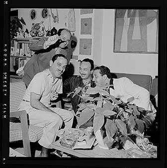 Four men are gathered in a living room, sitting and standing around with drinks and snacks, smiling and toasting in a social setting.