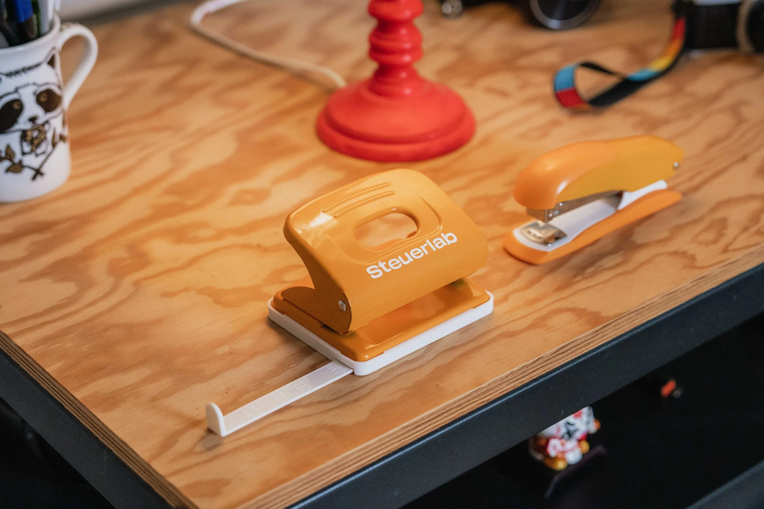 Ein orangefarbener Steuerlab-Klemmbrett auf einem Holzschreibtisch, in der Nähe befinden sich ein roter Kerzenständer, ein Tape-Meister, eine Tasse mit Tiermotiven und weiteres Büro- oder Bastelmaterial.