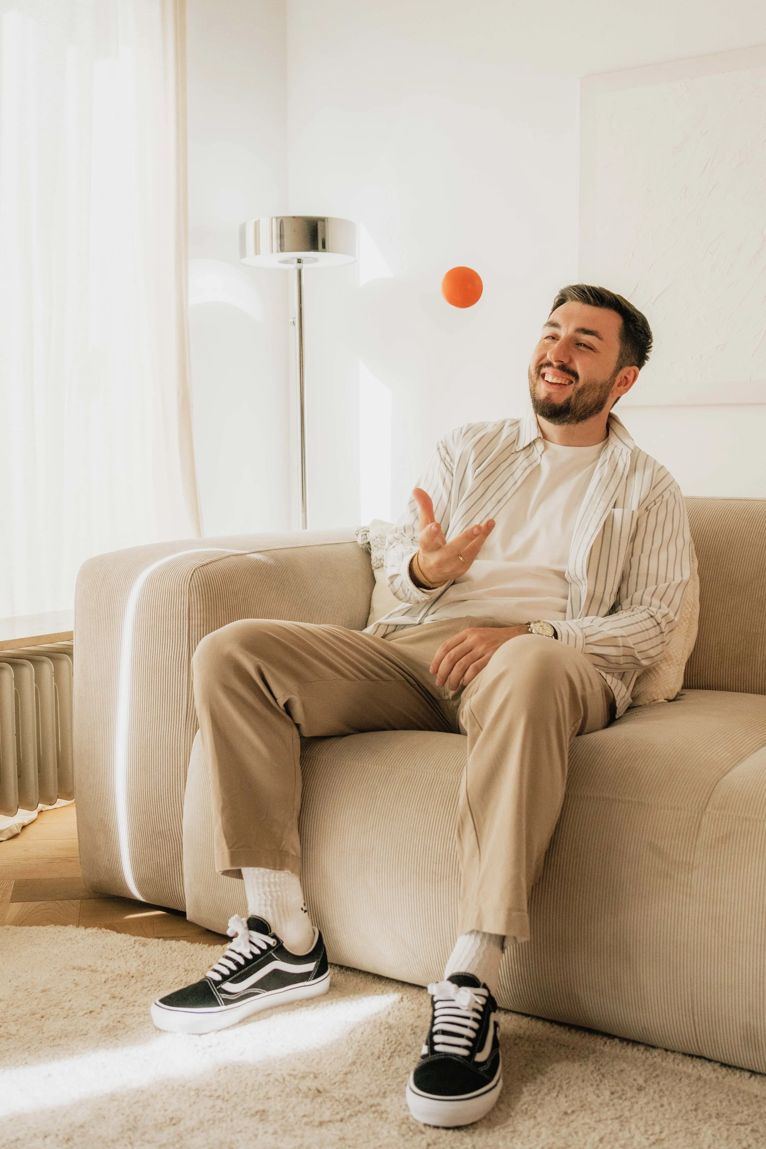 Ein Mann sitzt auf einem beigefarbenen Sofa, trägt beige Hosen, einen weißen T-Shirt, eine gestreifte Jacke, Vans Sneakers und lacht, während er einen roten Ball jongliert.