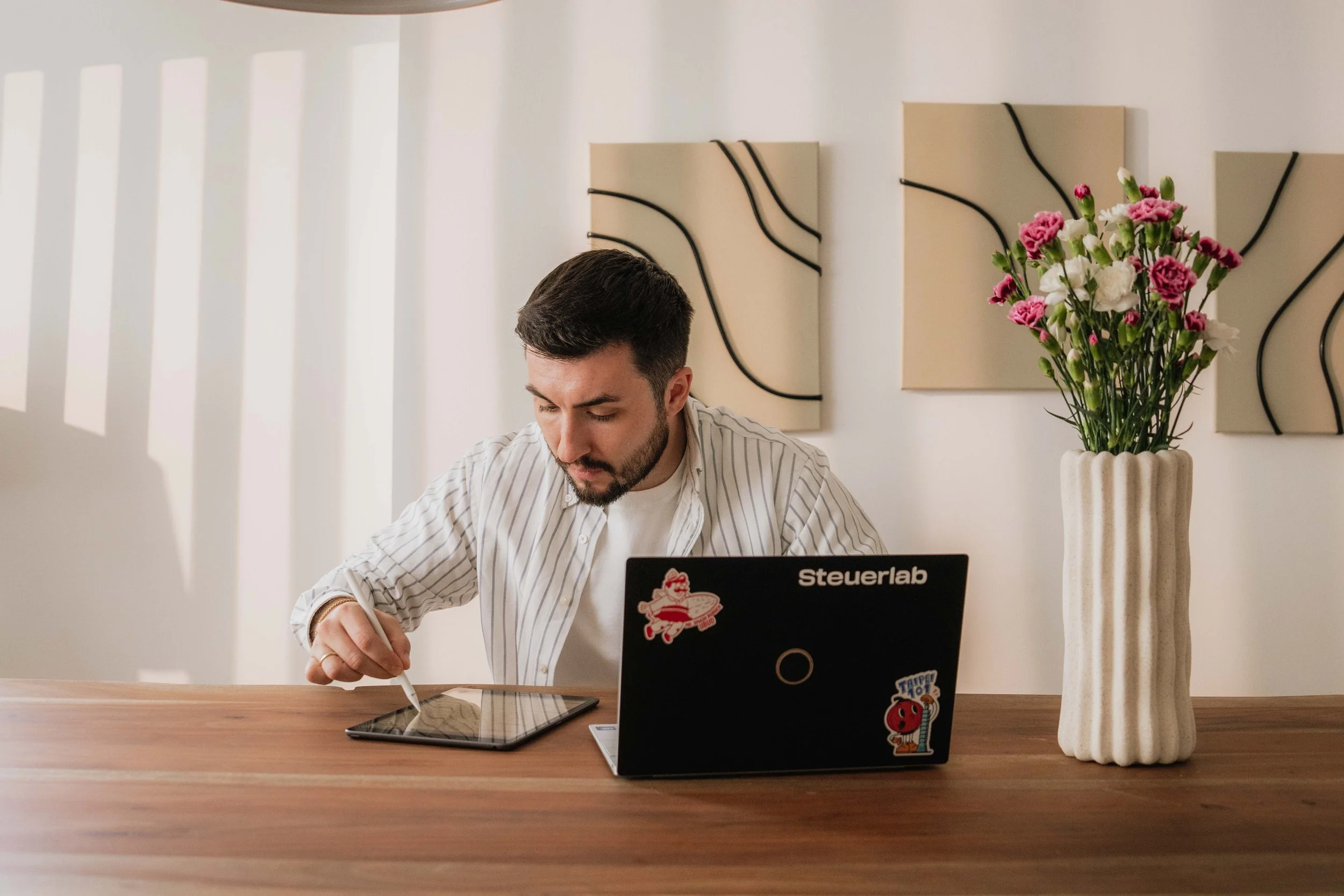 Ein Mann sitzt an einem Tisch mit einem Laptop, auf dem Sticker von Tappy Topp und dem Weihnachtsmann sind. Im Hintergrund ist eine Vase mit pink-weißen Blumen und abstrakten Wanddekorationen.