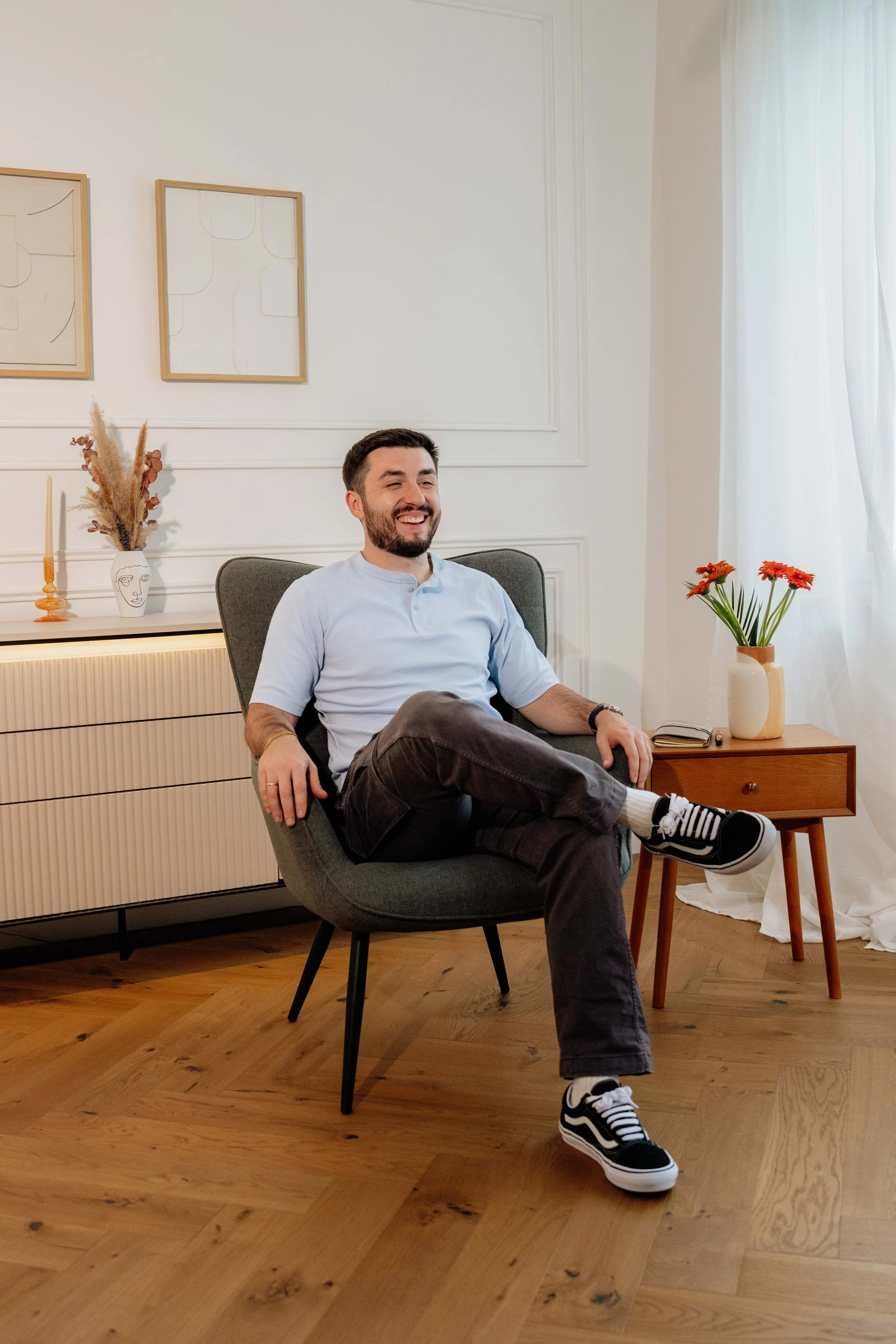 Ein lachender Mann sitzt entspannt in einem grauen Stuhl in einem hell eingerichteten Raum mit Holzfußboden. Neben ihm auf einem kleinen Tisch stehen eine weiße Vase mit orangefarbenen Blumen, ein Notizbuch und ein Kugelschreiber.
