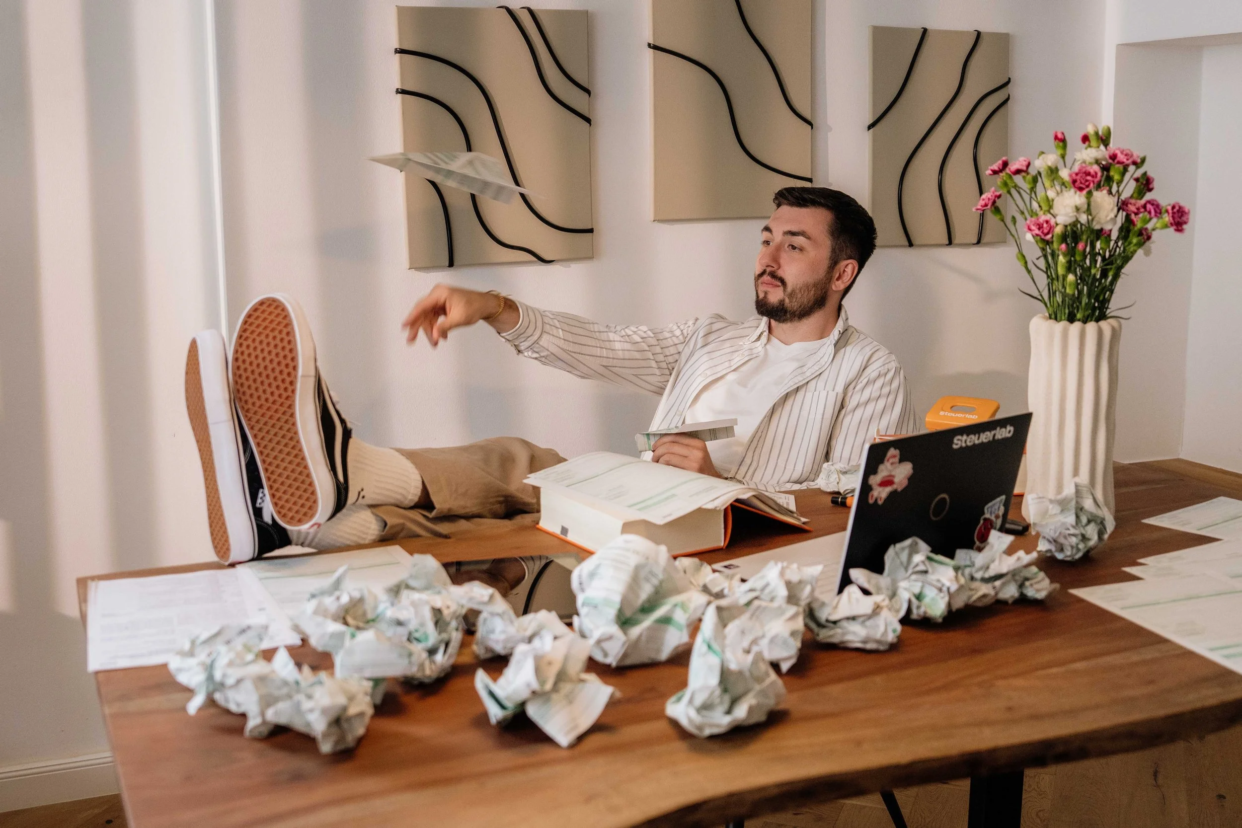 Ein Mann sitzt entspannt an einem Tisch, seine Füße liegen auf dem Tisch. Der Tisch ist unordentlich mit Papieren, zerknülltem Papier und Büchern bedeckt. Im Hintergrund sind drei moderne, abstrakte Wanddekorationen und eine große Vase mit bunten Blumen zu sehen.