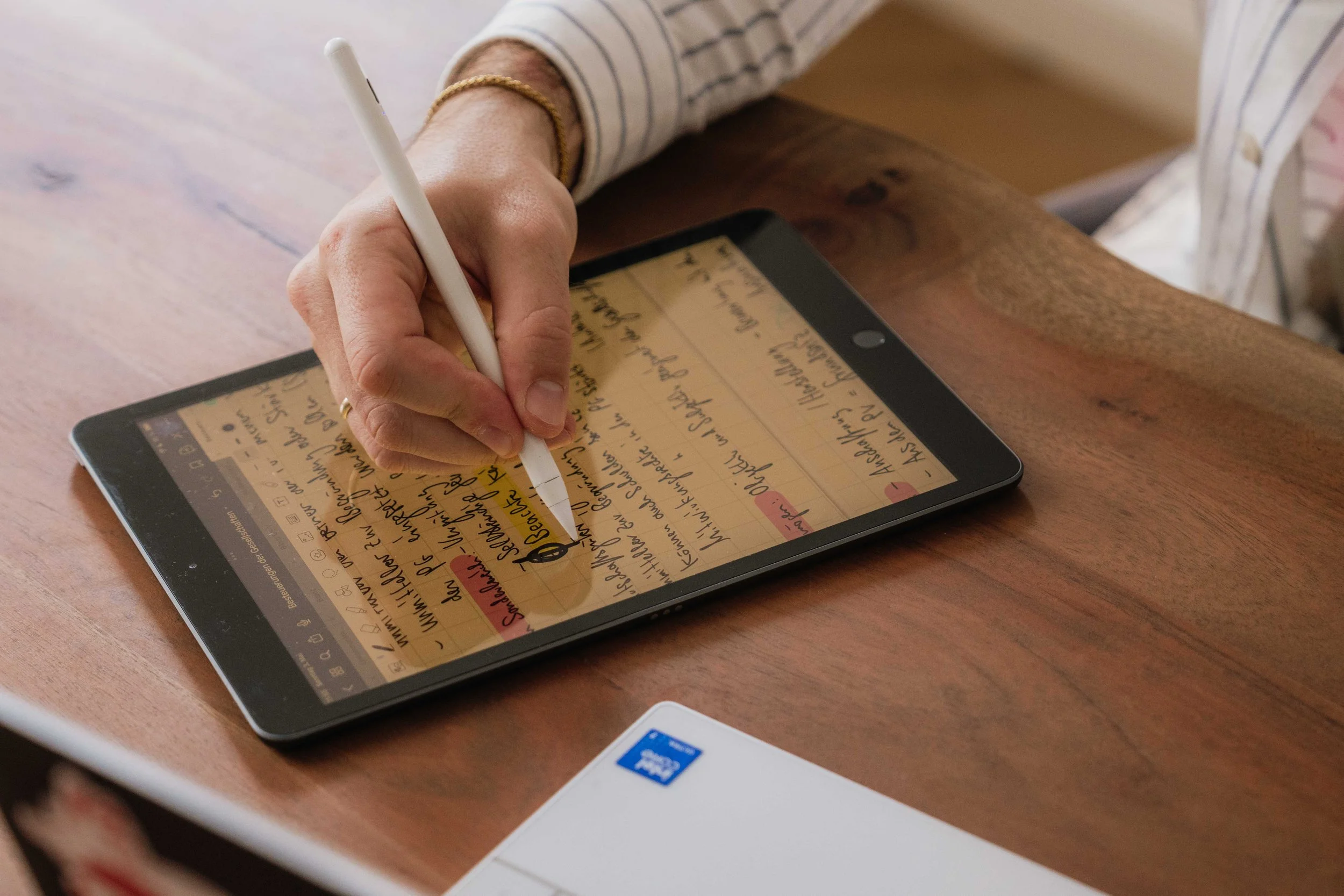 Hand schreibt mit einem weißen Stift in ein digitales Notizbuch auf einem Tablet, das auf einem Holztisch liegt.