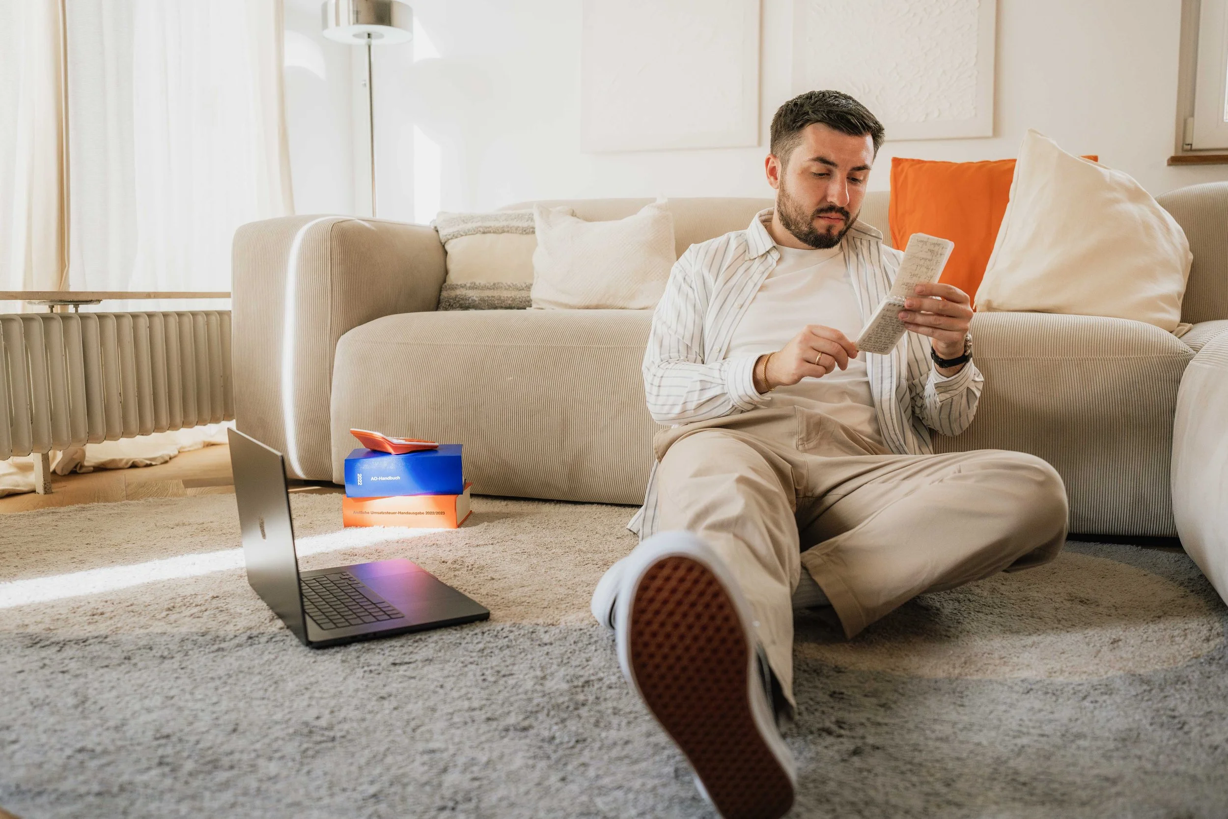 Ein Mann sitzt auf dem Boden vor einem Sofa, liest in einem Notizbuch und hat einen Laptop, mehrere Bücher und Notizen um ihn herum. Helle, freundliche Wohnzimmeratmosphäre.