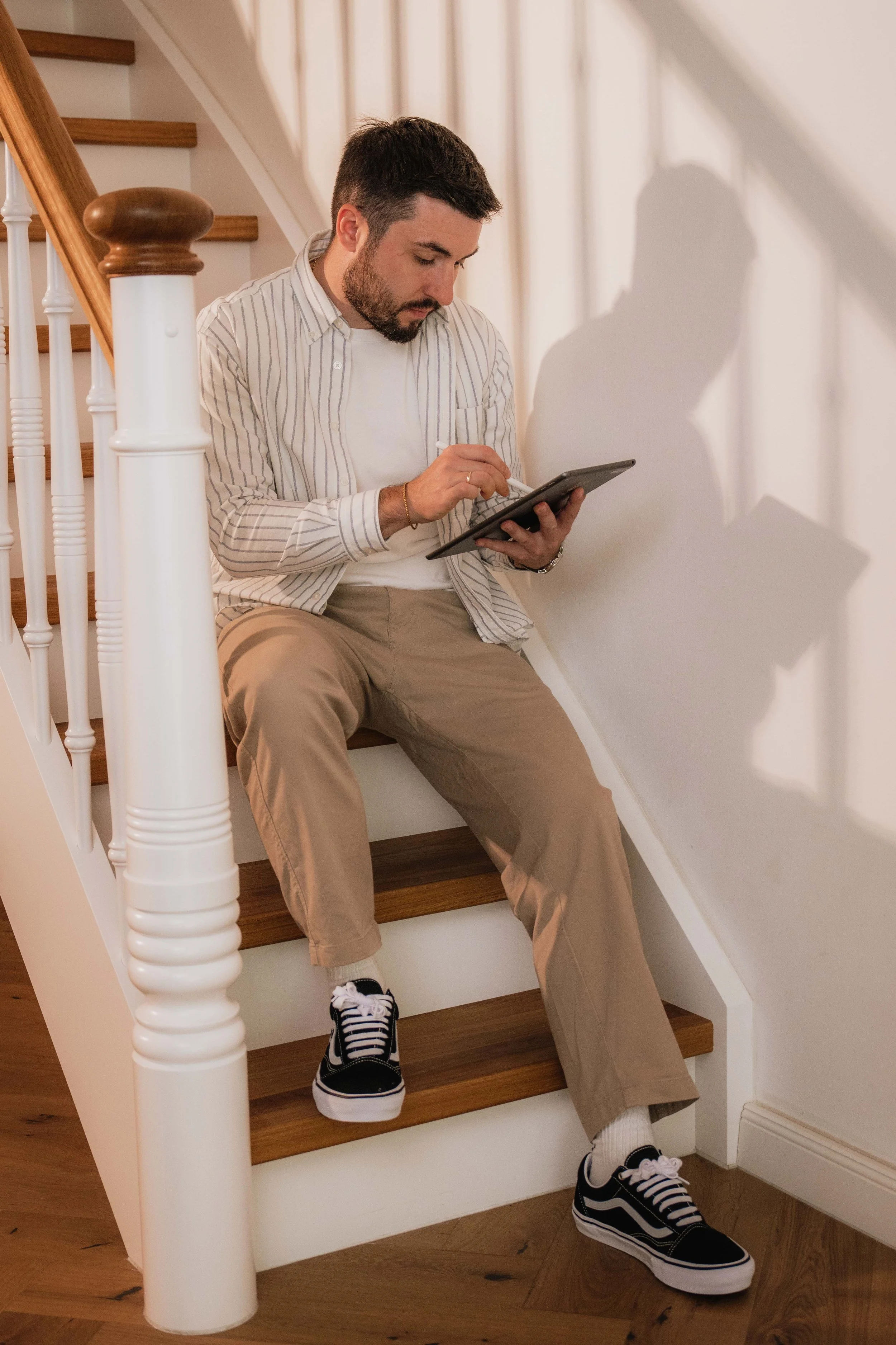 Ein Mann sitzt auf einer Treppe neben einer weißen Wand, trägt eine gestreifte Hemd, beige Hosen und schwarze Turnschuhe und schaut auf ein Tablet.