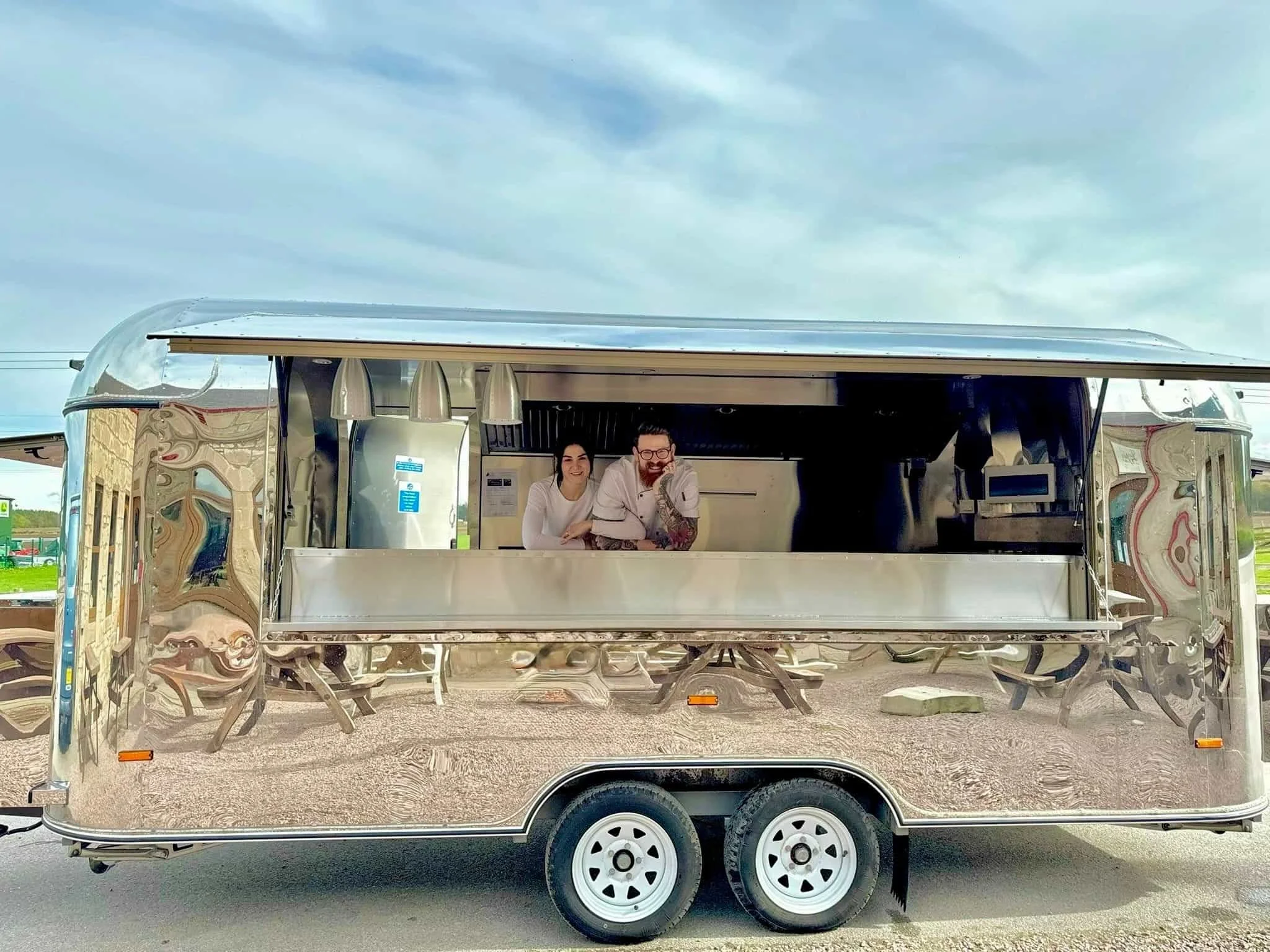 Two people inside a shiny, reflective food trailer, smiling at the camera, standing behind an empty counter, with a cloudy sky in the background.